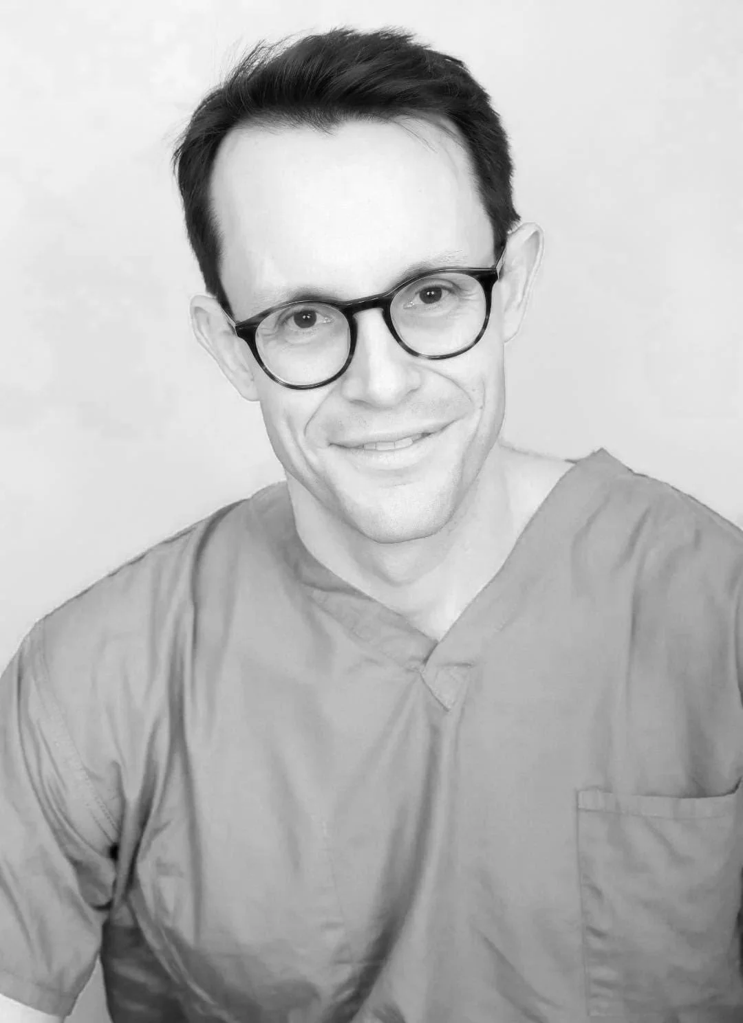Black and white photo of a smiling man wearing round glasses and scrubs.