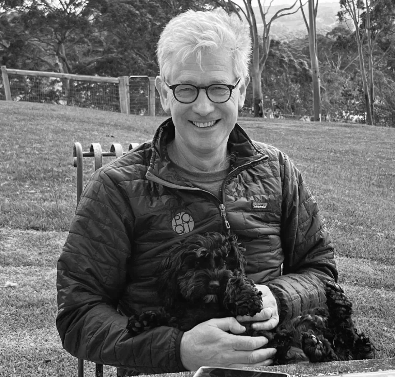 A man with glasses smiling and sitting outdoors on a chair, holding a small black puppy in his arms. He is wearing a quilted jacket, and there are trees and a fence in the background.