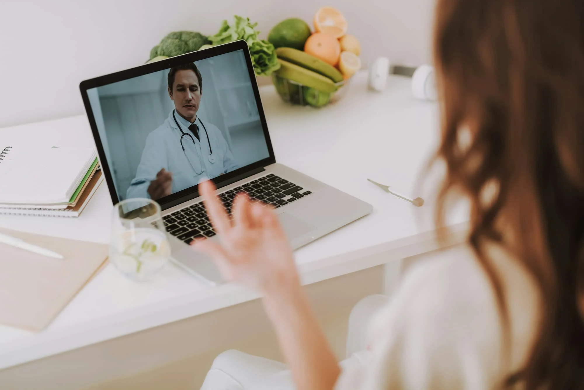 A woman is having a video call with a male doctor on her laptop. The scene is set on a white table with a glass of water, a notebook, and a spoon. Behind her, there is a fruit bowl with bananas, apples, and lemons, and some green vegetables. The woman has curly brown hair, and her hand is raised with her index finger pointing or gesturing.
