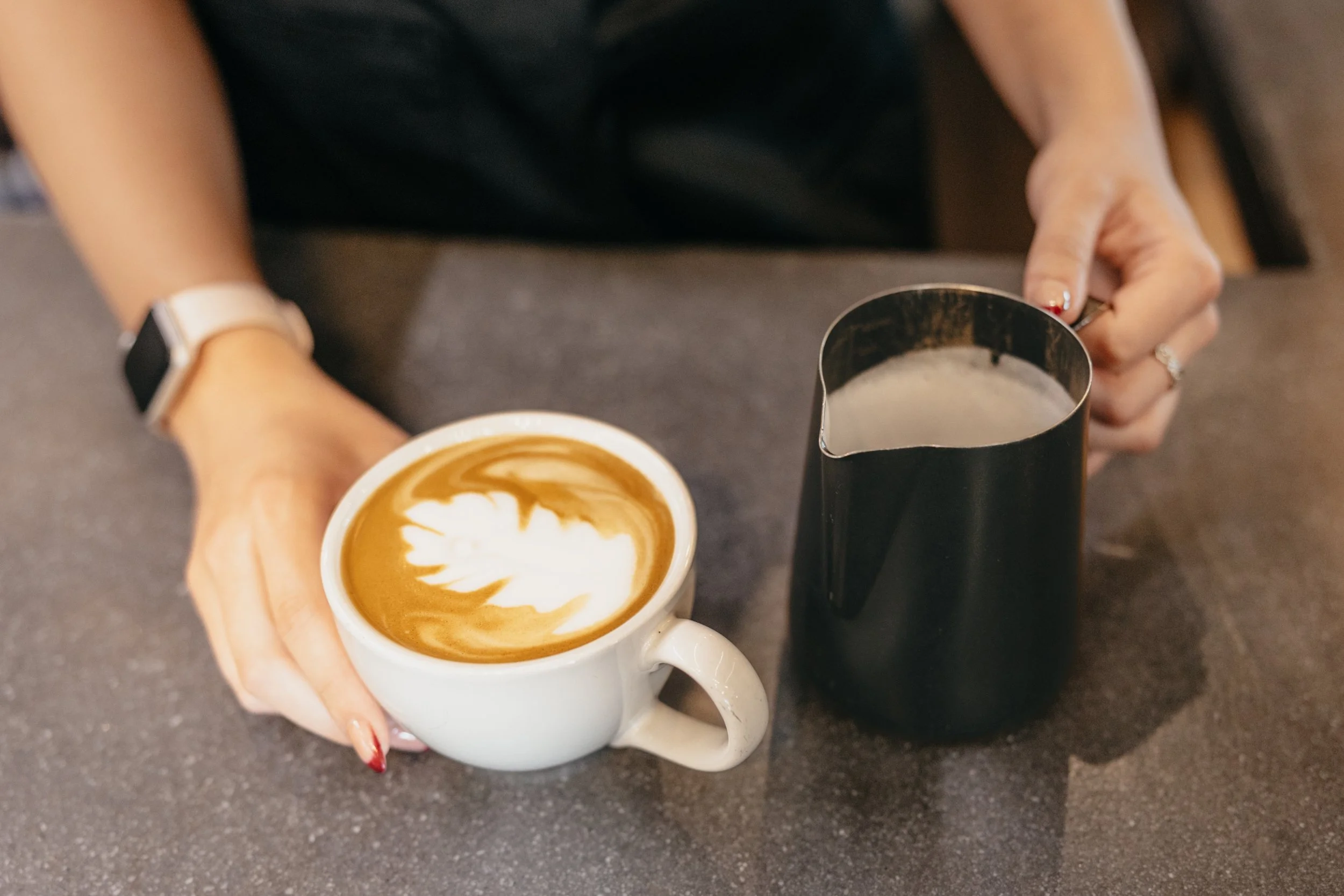 A person holding a white ceramic cup of latte with latte art, along with a black milk pitcher on a gray countertop.