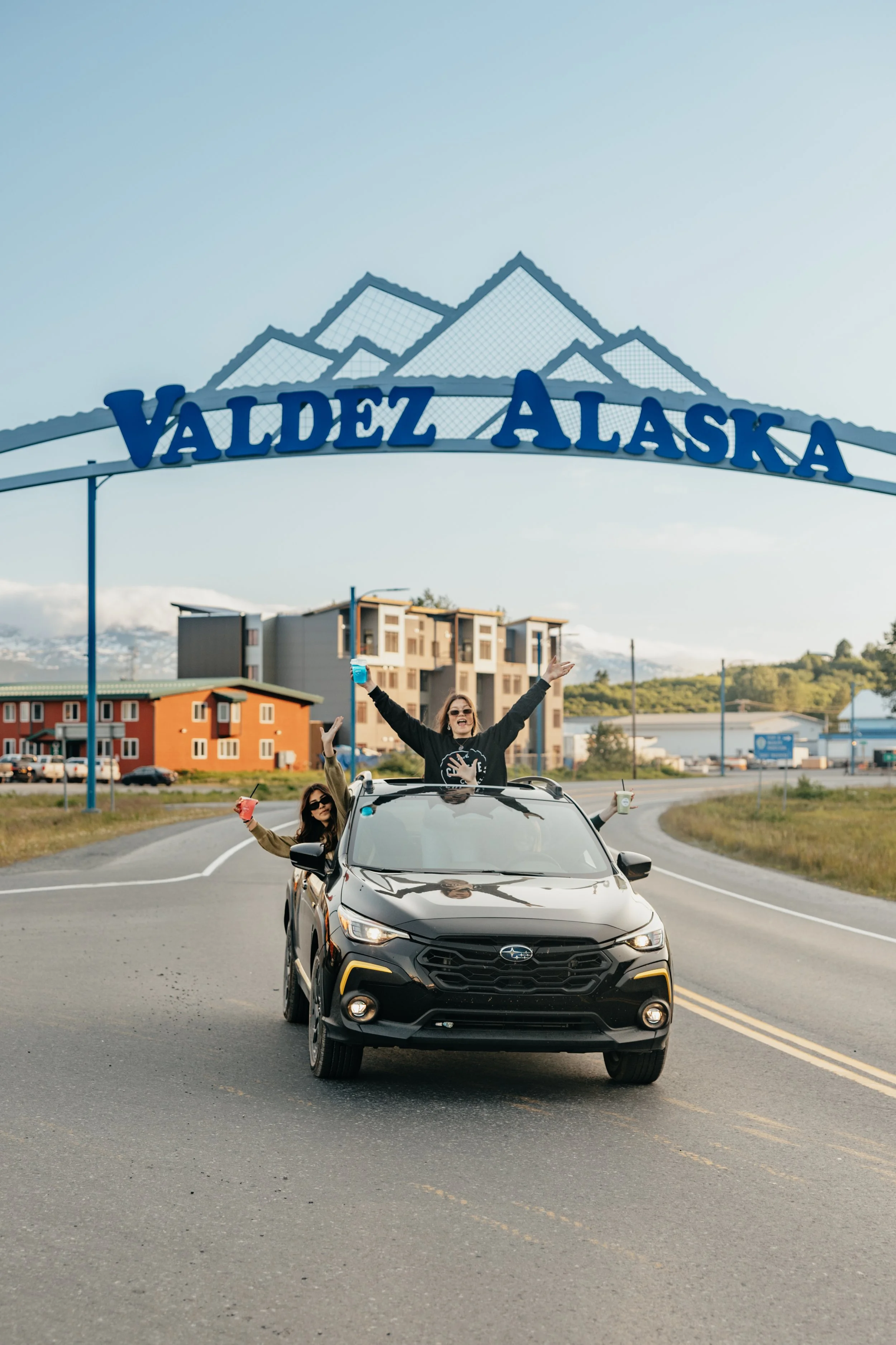 People in a black car celebrating under a sign that says 'Valdez Alaska' on a road.