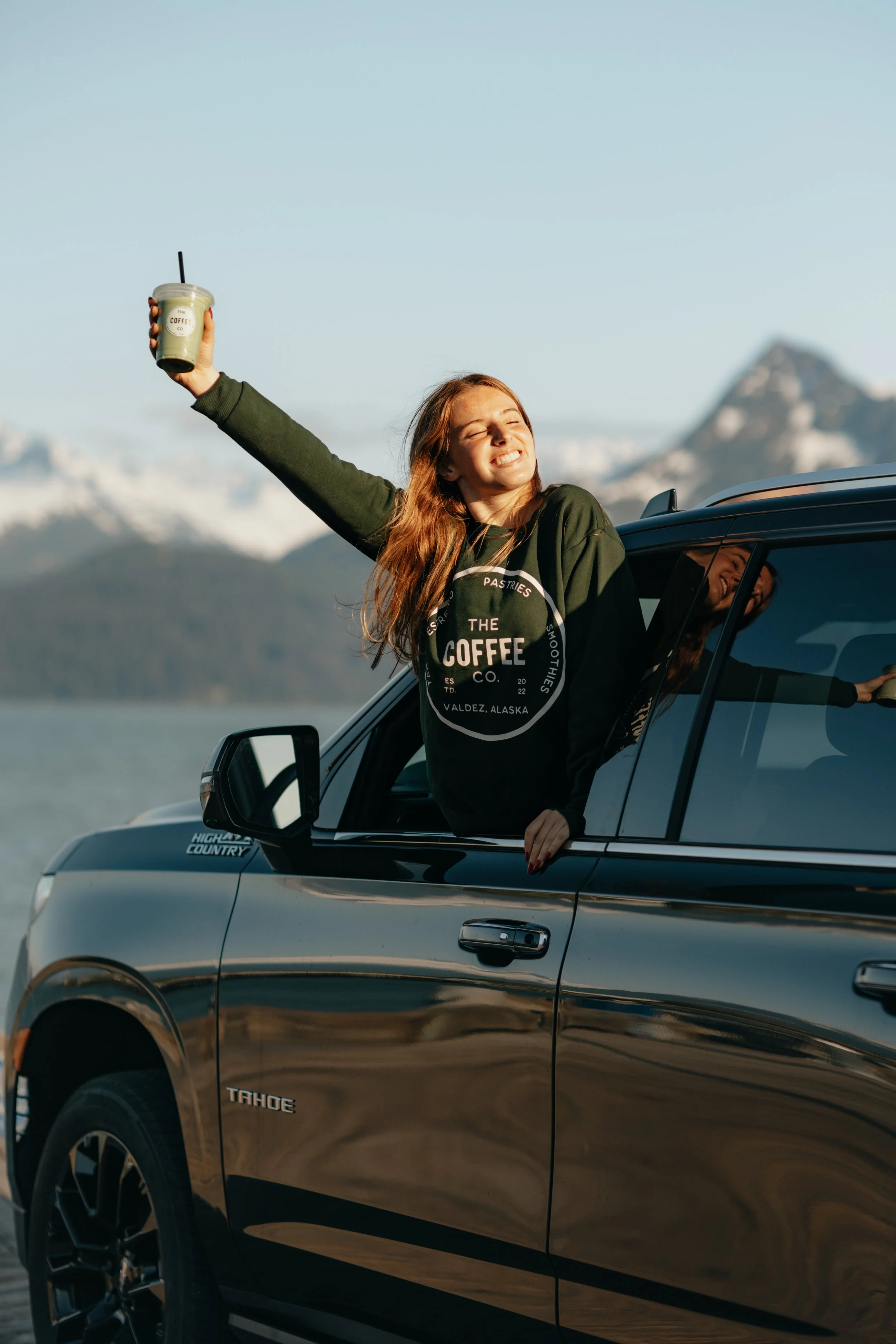 A woman with red hair leaning out of a black SUV window, smiling with eyes closed, holding a plastic cup with a straw, outdoor landscape with snow-capped mountains in the background.