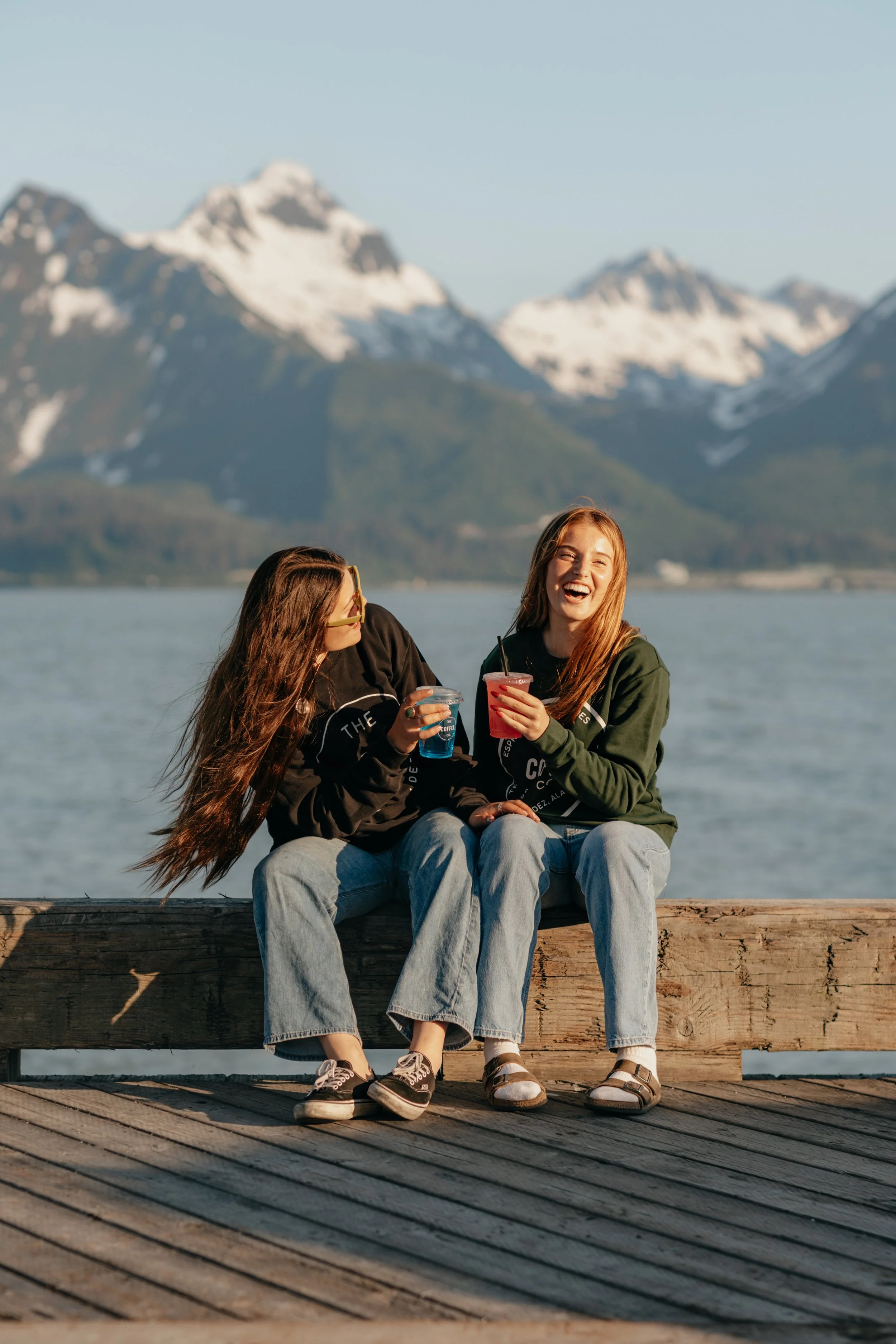 Two young women sitting on a wooden dock by a lake, enjoying drinks, with snow-capped mountains in the background.