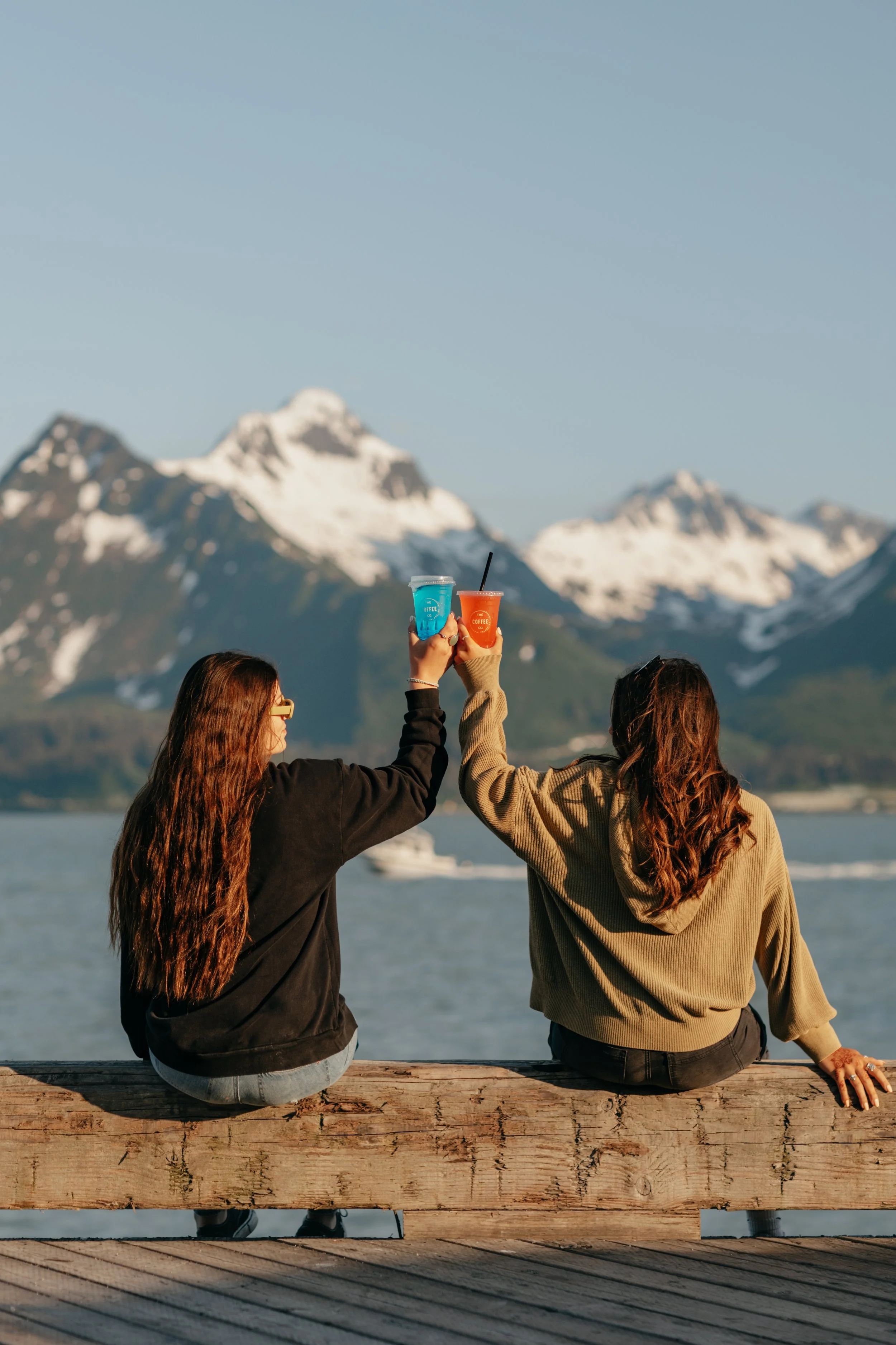 Two women sit on a wooden dock, raising colorful drinks towards snow-capped mountains, with a body of water and clear blue sky in the background.