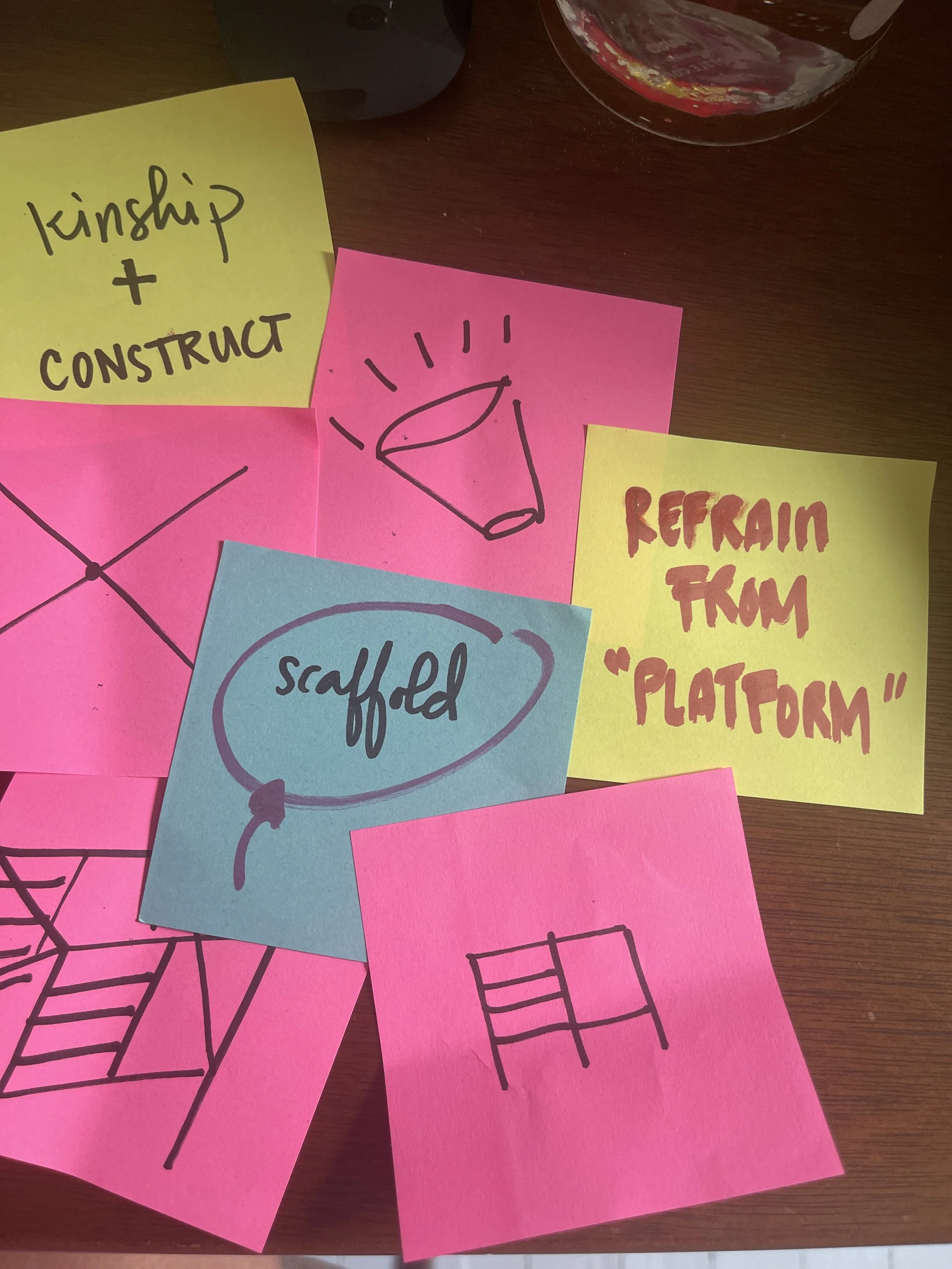 An ideation and conceptual design session: Hand-drawn sketches on colorful sticky notes arranged on a table, including diagrams of a microphone, a ladder, and handwritten notes reading "KINSHIP + CONSTRUCT" and "REFRAIN FROM PLATFORM''.