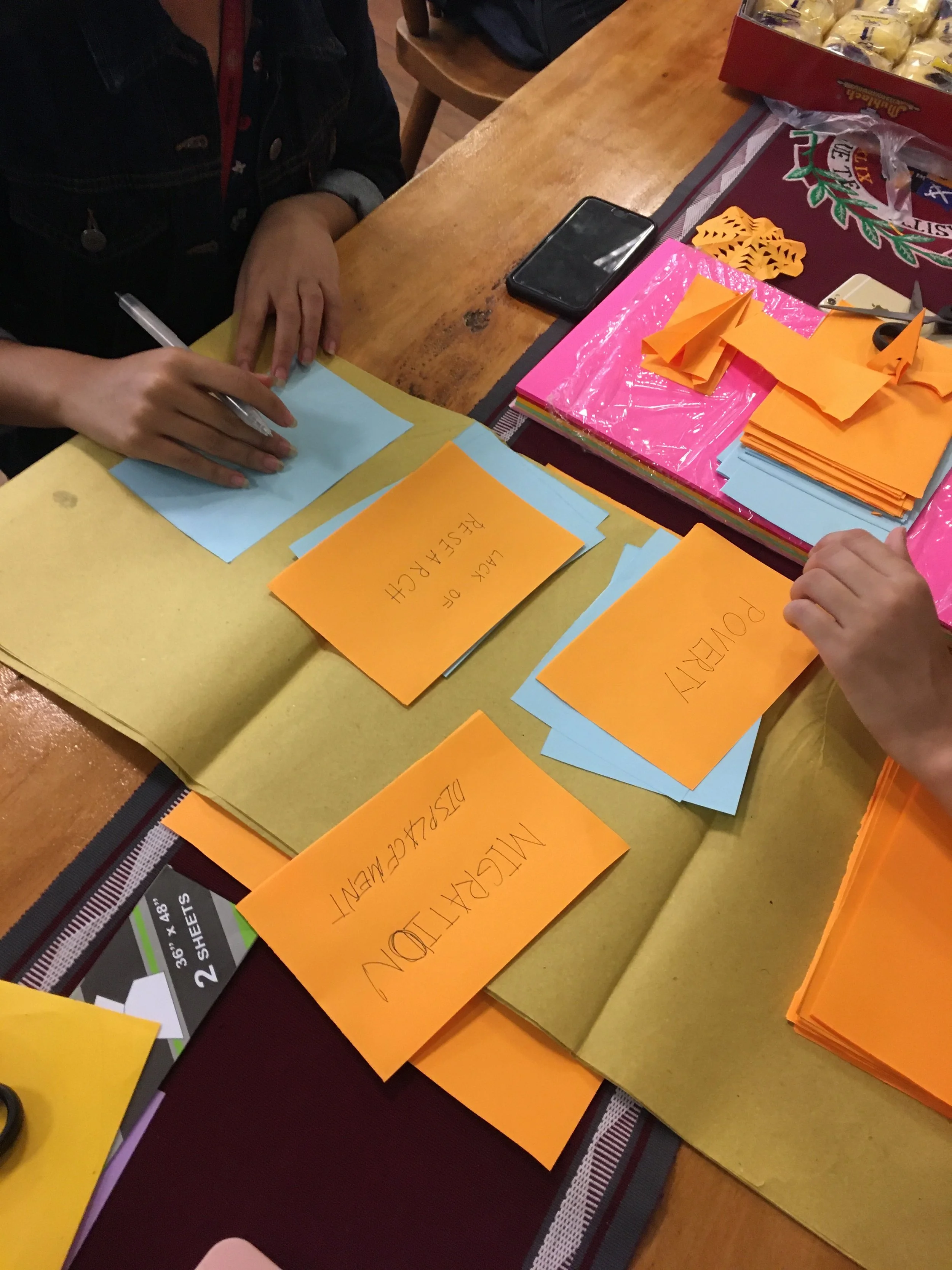 A close-up view of a sense-making session: A person writing on blue paper surrounded by scattered colored cards, a smartphone, orange paper, scissors, and stickers on a wooden table.