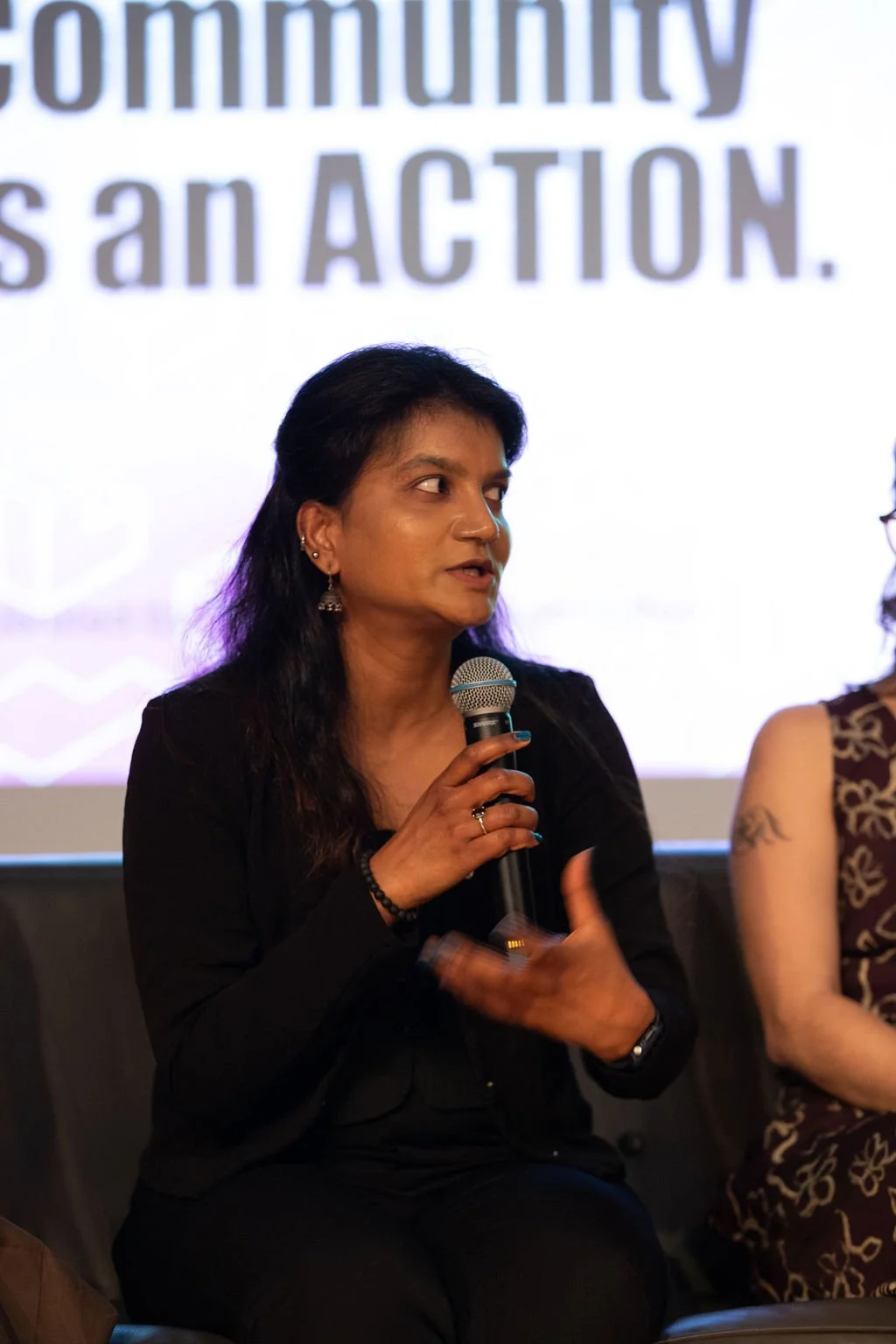 Swathi Gangadhar speaking into a microphone at a panel event for social impact design. Background display text with the words 'community is an ACTION.'
