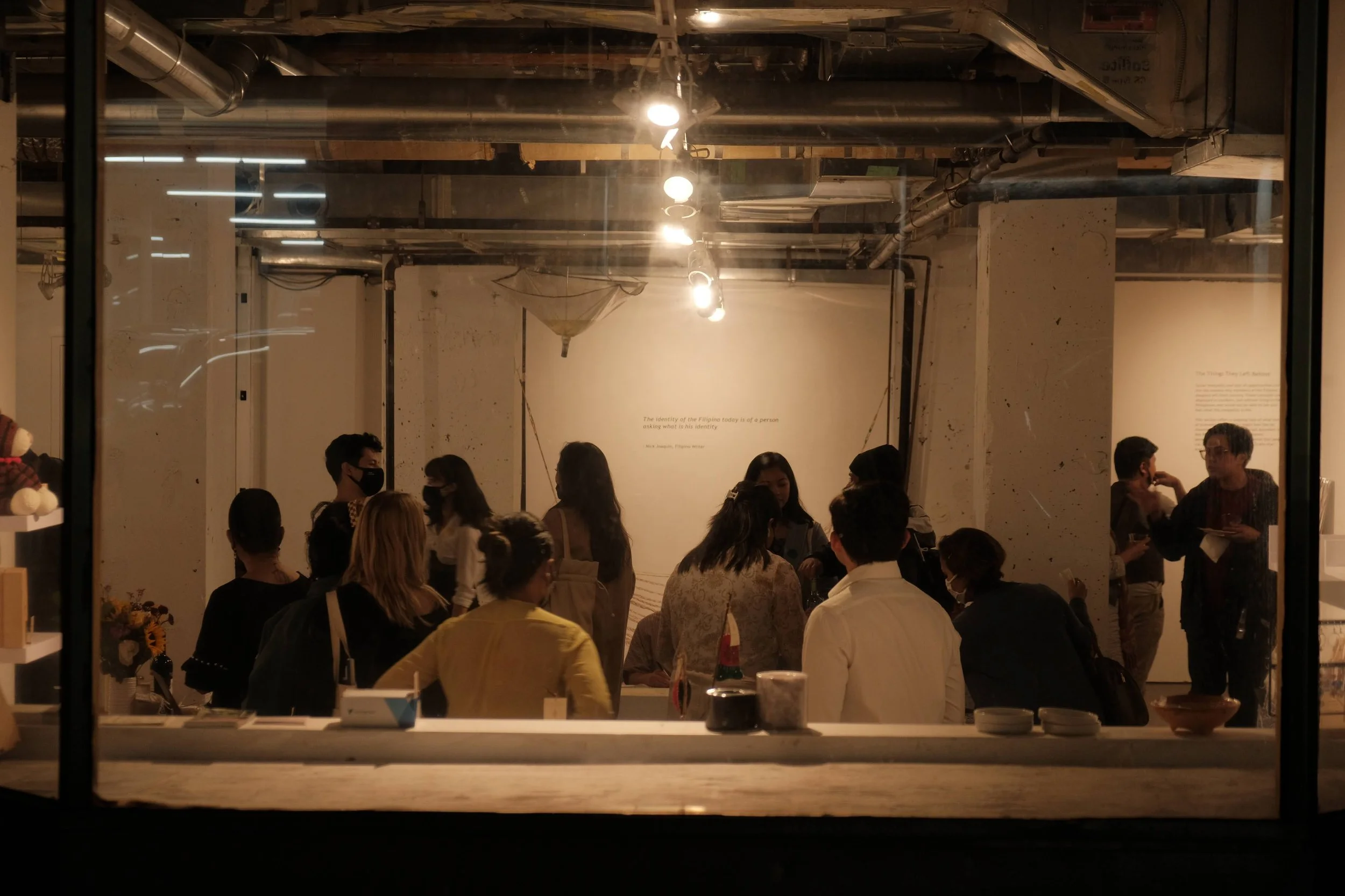 Photo of Bagay at Buhay pop-up gallery in November 2021. People gathered in an art gallery, seen through a window, with artworks and spotlights visible inside.