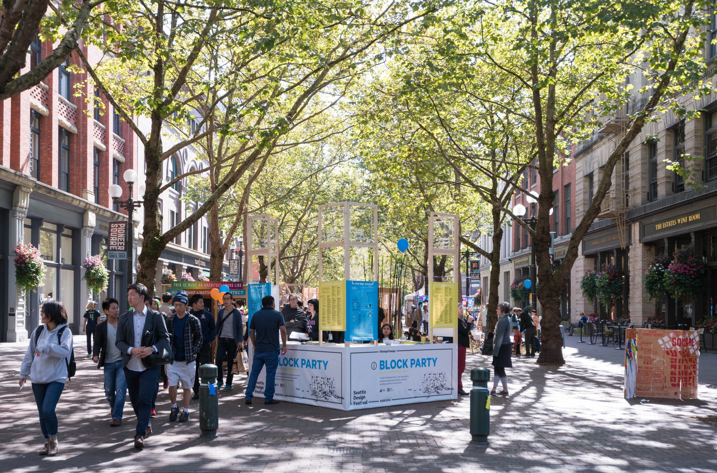 Outdoor block party with people, trees, and buildings in a city street, featuring a kiosk with banners for a design festival.