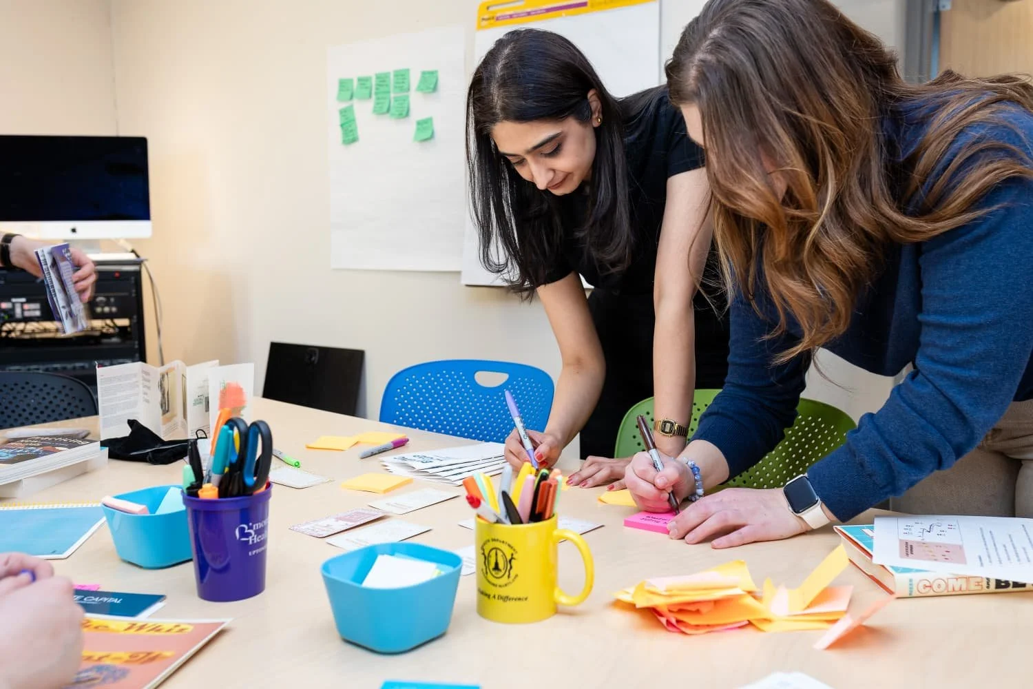 A collaborative brainstorming session: Two women work together at a table covered with sticky notes and office supplies, with a whiteboard.