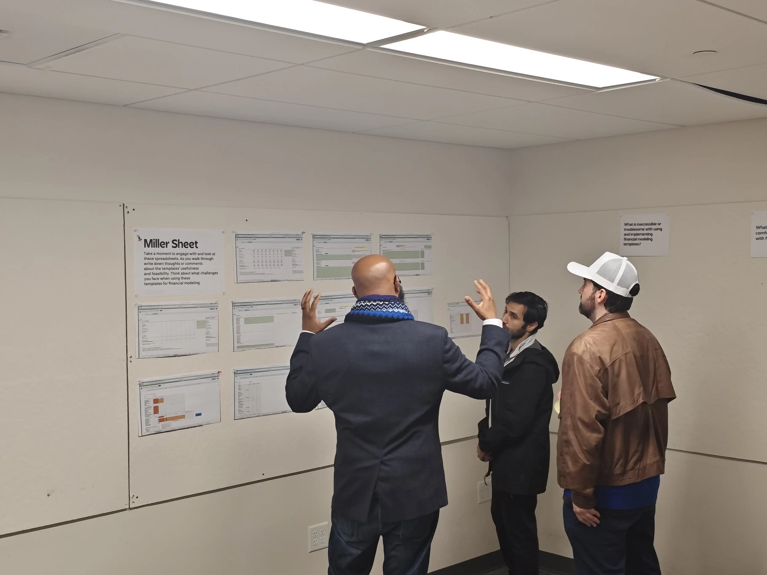 Four people standing and engaging in a discussion in front of a wall with printed spreadsheets and charts, with ceiling panels and lights visible above.