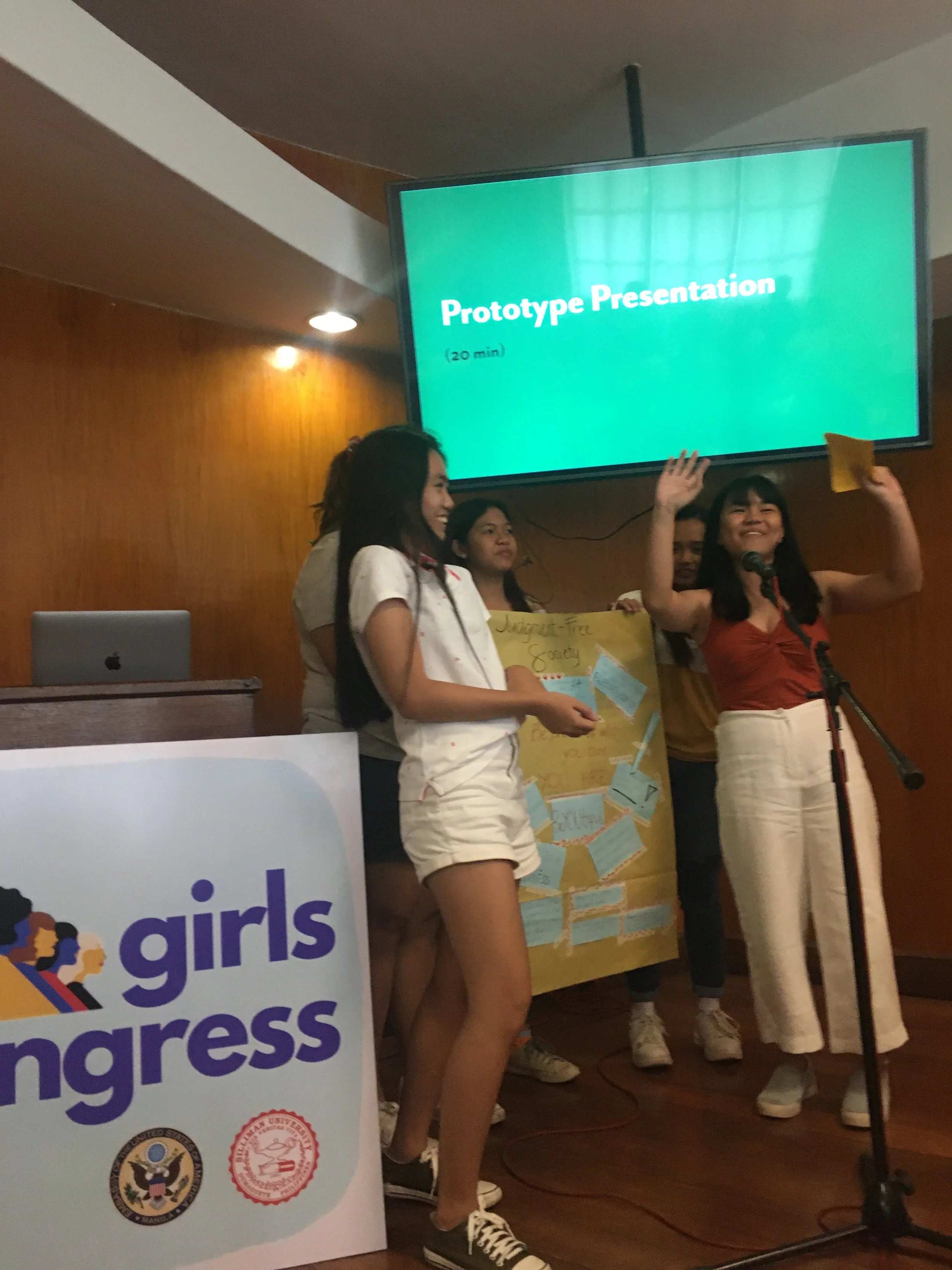 Group of girls presenting on stage at Girls Congress event, with a woman speaking into a microphone, holding yellow paper, and a large screen displaying 'Prototype Presentation' in the background.