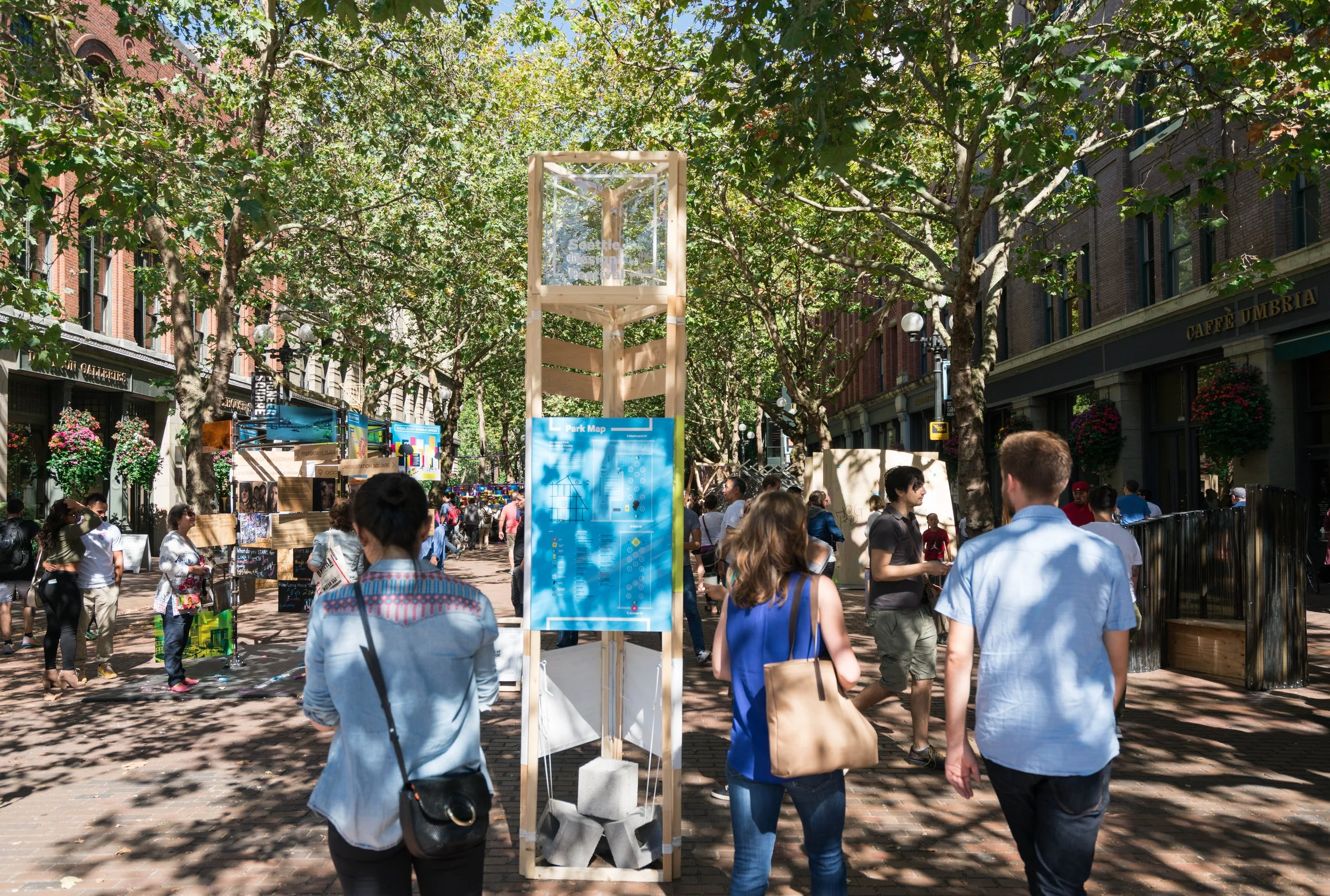 Busy pedestrian street with trees, informational display, people walking, shops with hanging plants, and a park map under clear blue sky.