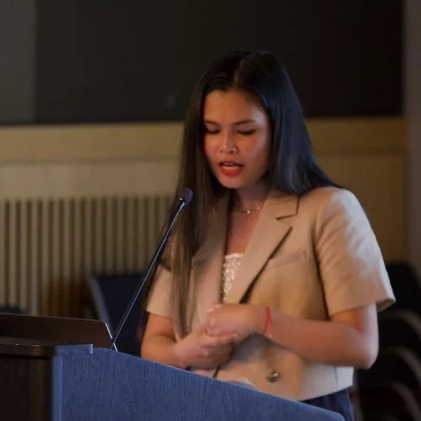 Ana Mengote Baluca speaking at a social design event podium with a microphone, wearing a beige blazer and a white top.