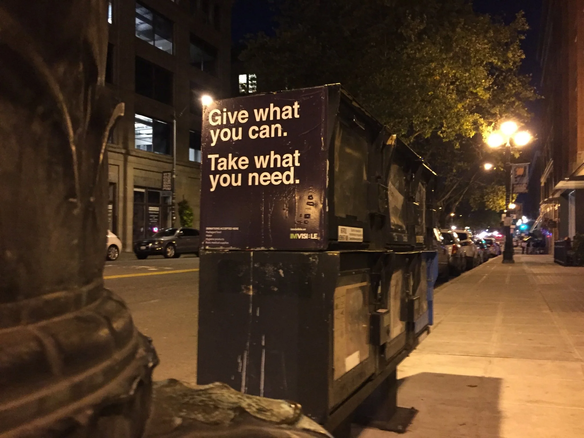 A street scene at night with a sign that reads "Give what you can. Take what you need." The street is lined with parked cars, overhead lights, and trees.