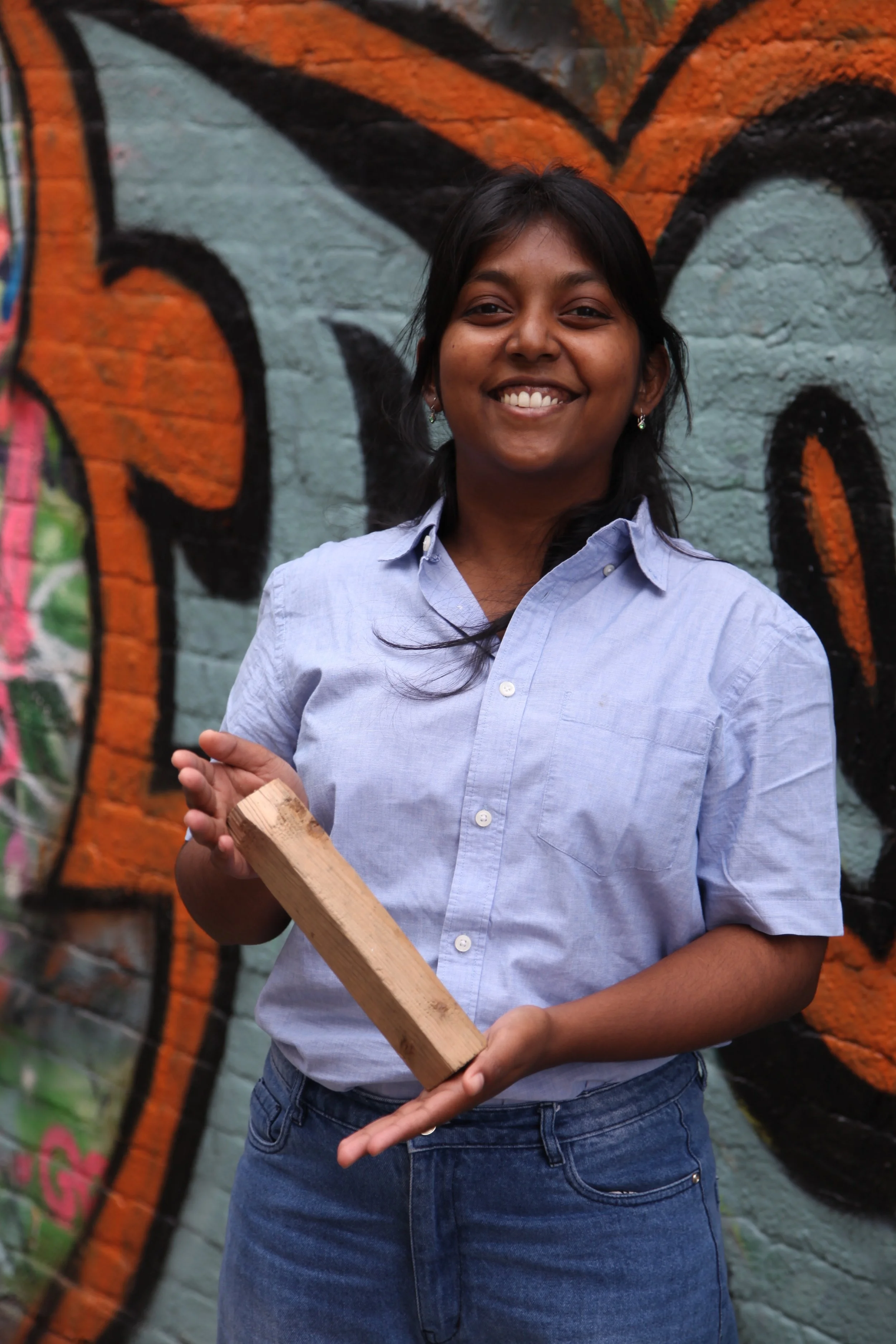 Pallavi Mambillil smiling holding a wooden block she found beautiful in front of colorful graffiti wall.