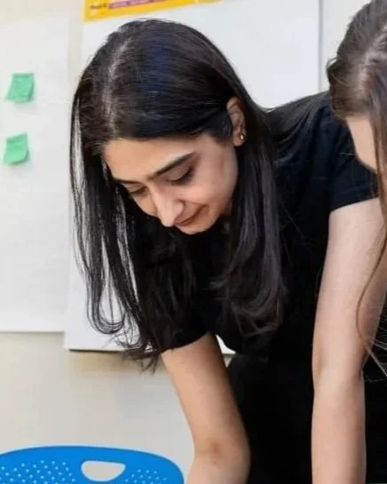 A woman with long dark hair looking down, bending over a table in a classroom setting.