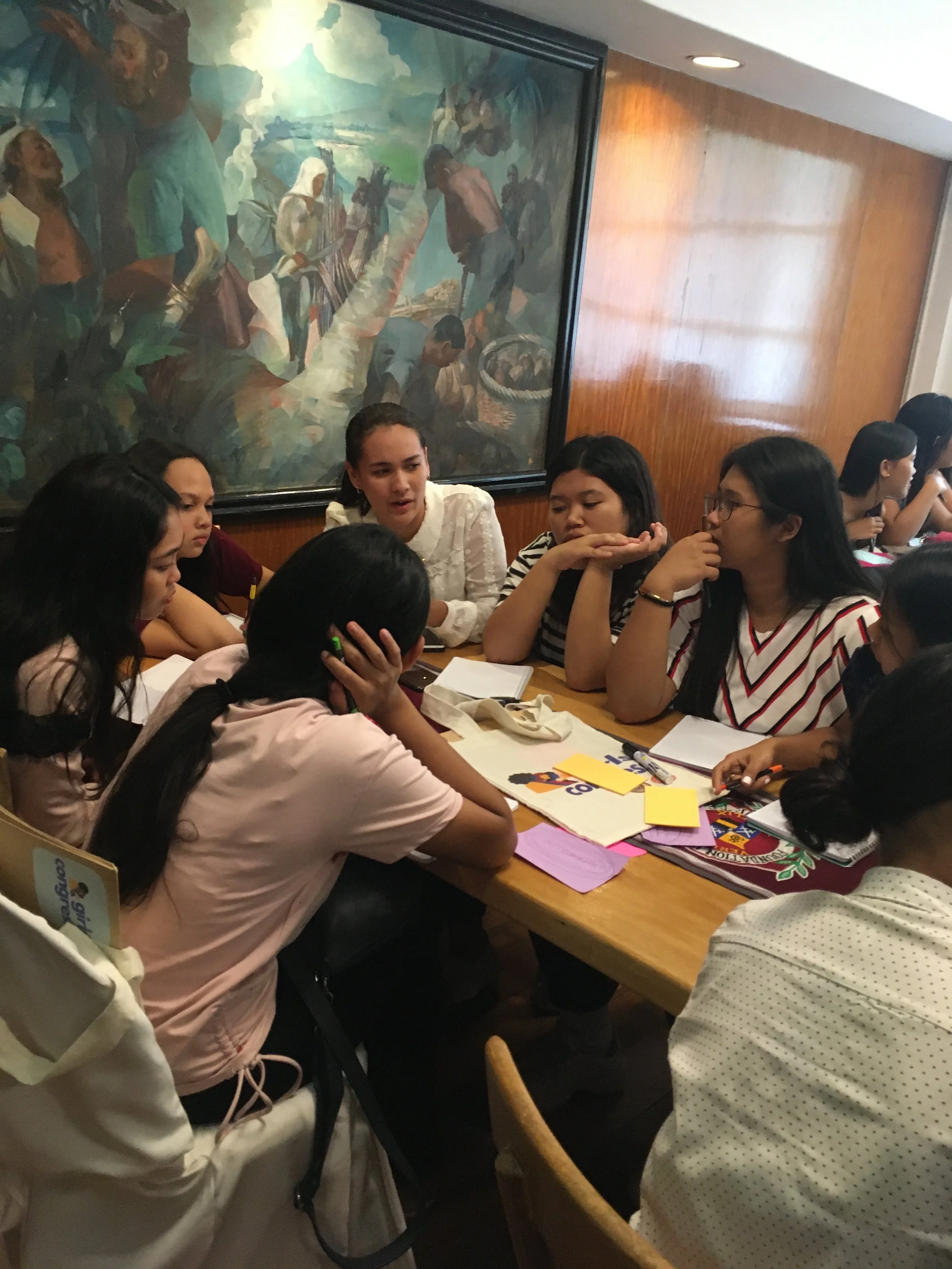 A group of young women gathered around a table engaged in a discussion, with notebooks and papers in front of them, in a room with a large painting on the wall.