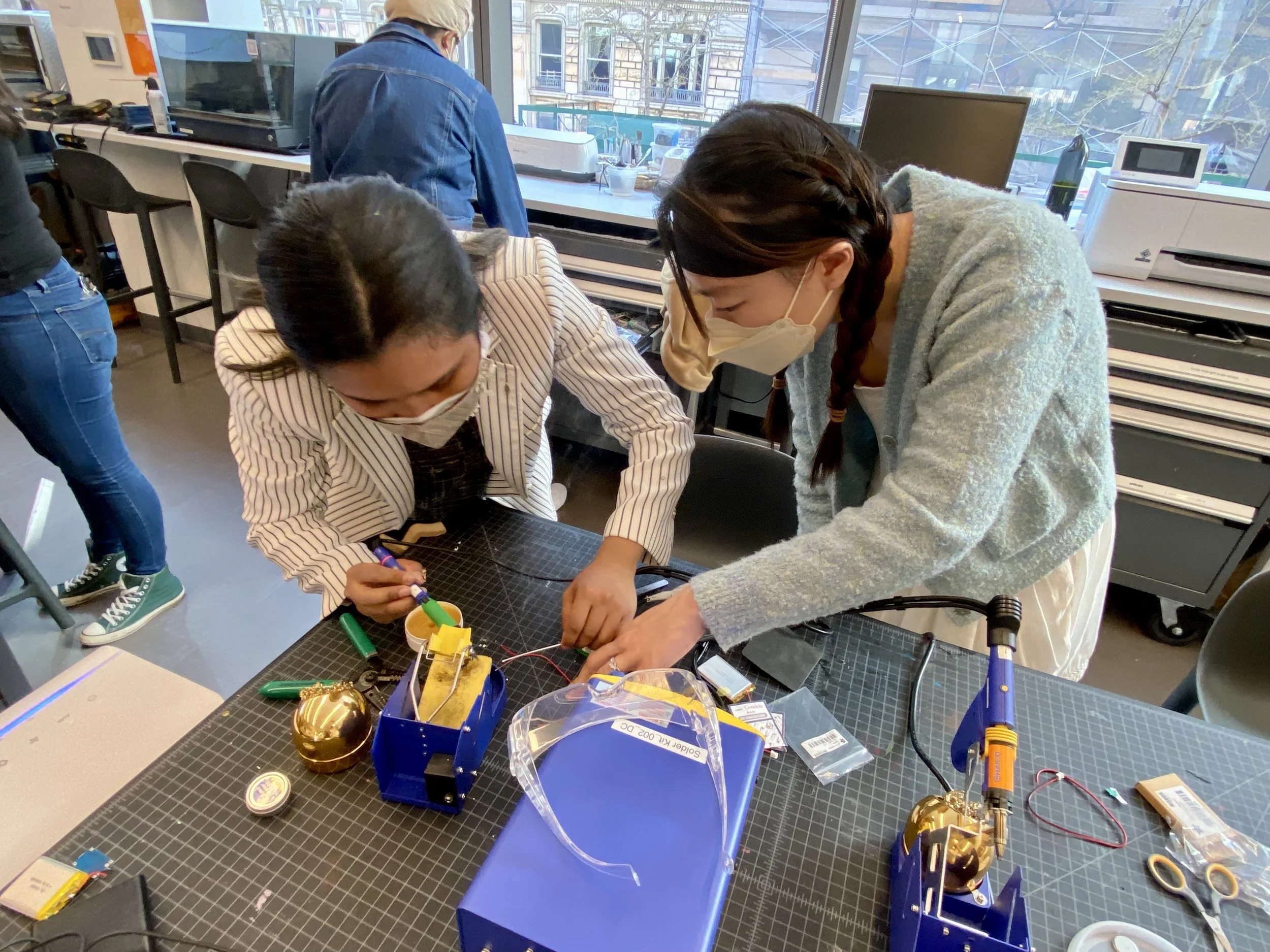 A product design session: Two women wearing masks collaborate on an electronics prototype at a workstation filled with tools and hardware components, with one leaning over the table while the other adjusts a component.