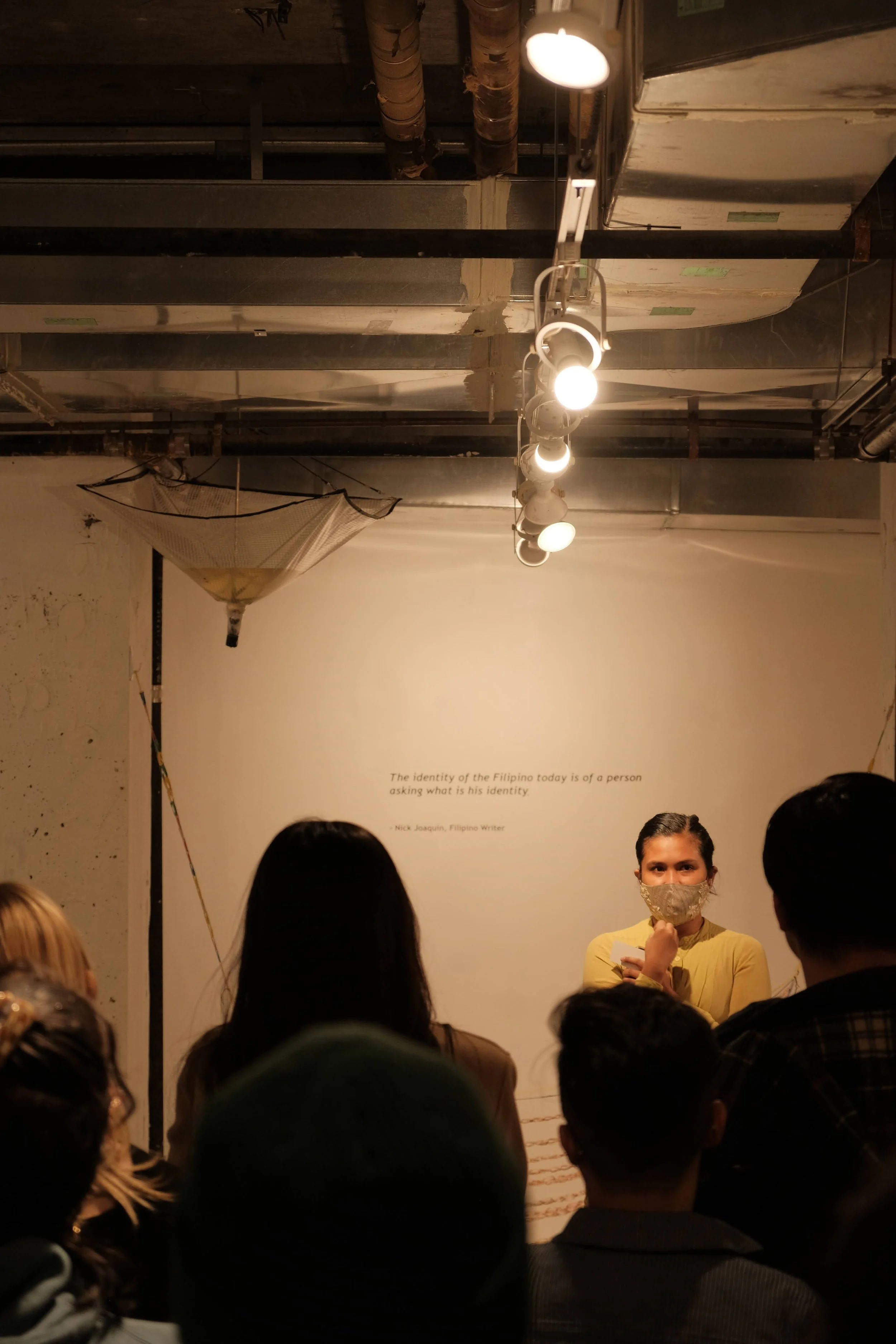 A woman wearing a face mask standing in front of a white wall with a quote. Several people are gathered around her, listening. The ceiling has exposed ductwork and a track lighting system.