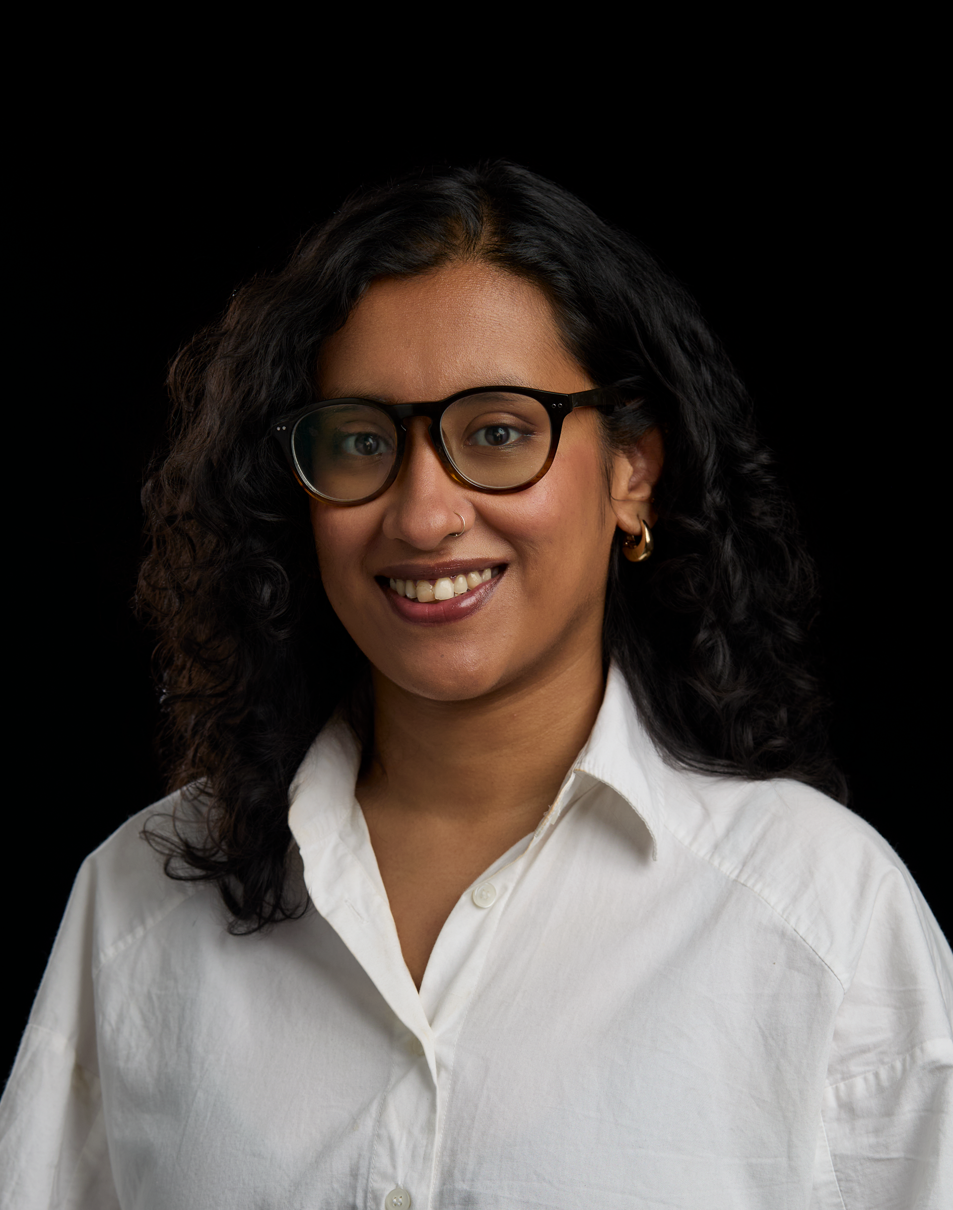 Close-up of a smiling woman with curly black hair, wearing black glasses and gold hoop earrings, dressed in a white button-up shirt, against a black background.