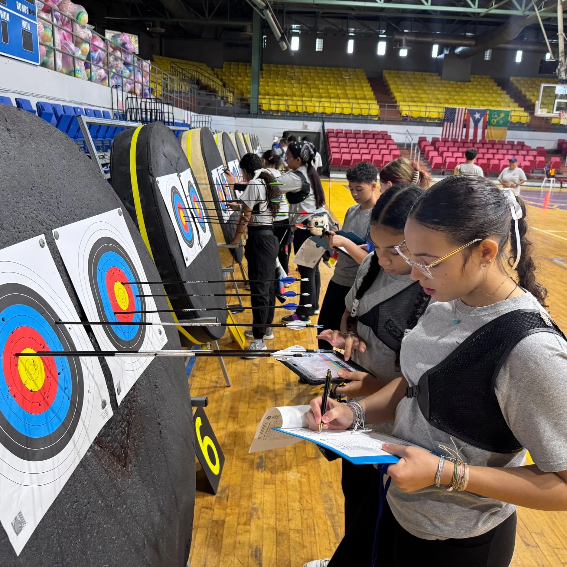 Group of archery athletes shooting arrows at archery targets in an indoor gymnasium, some taking notes and scores, then  aiming their bows, with seating and flags visible in the background. NFAA Indoor Target League. Puerto Rico Archery League.