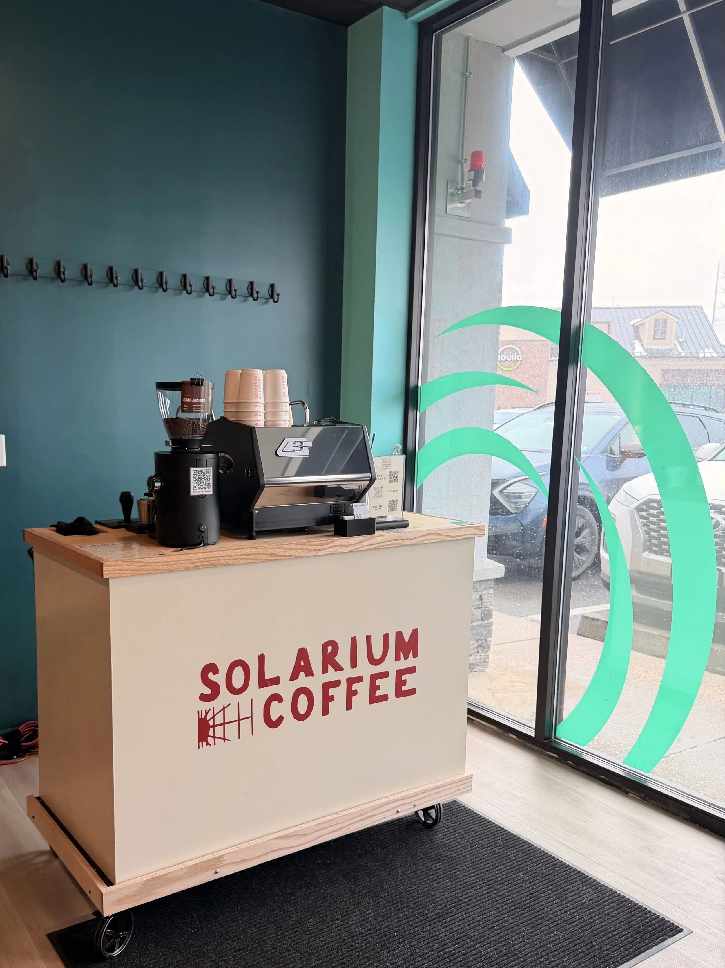 Inside a coffee shop with a small service counter labeled Solarium Coffee, featuring a commercial espresso machine, cups, and a coffee grinder. Large window with teal designs, parked cars outside.