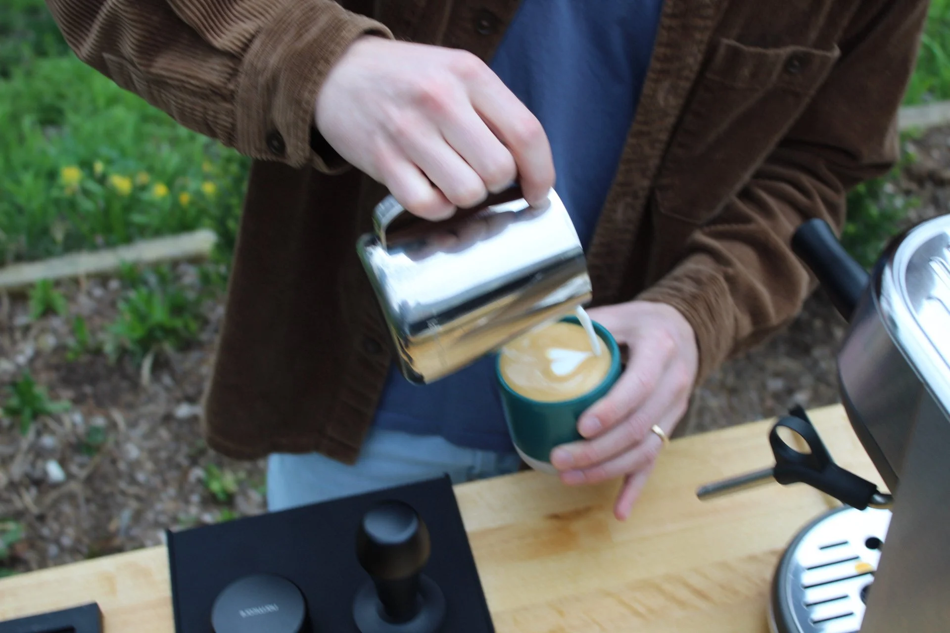 A person pouring steamed milk from a metal pitcher into a latte in a cup, creating latte art with a heart shape. The person is wearing a brown jacket and a ring on their finger, and they are standing outside near a wooden table with a coffee machine nearby.