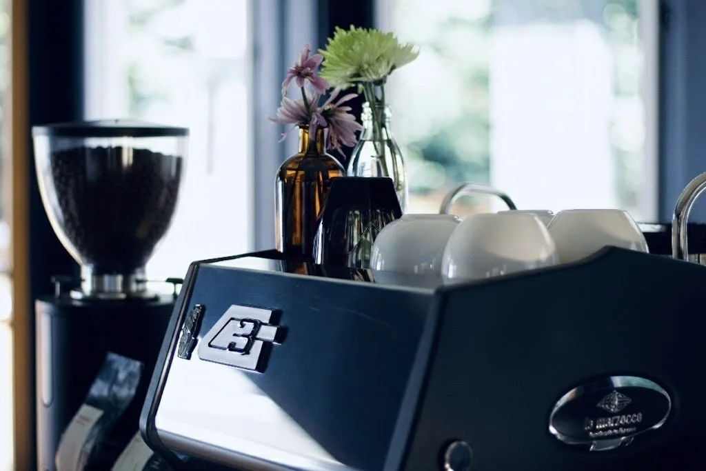 Coffee grinder, vases with flowers, coffee cups, and coffee machine on kitchen counter in front of a window.
