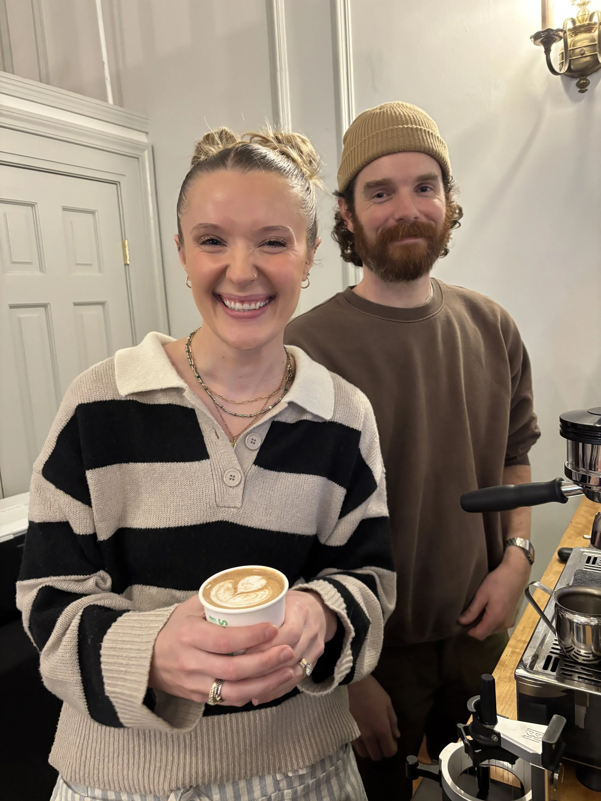 A smiling woman holding a cup of coffee with latte art, standing next to a man with a beard and a tan beanie, in what appears to be a coffee shop or cafe.