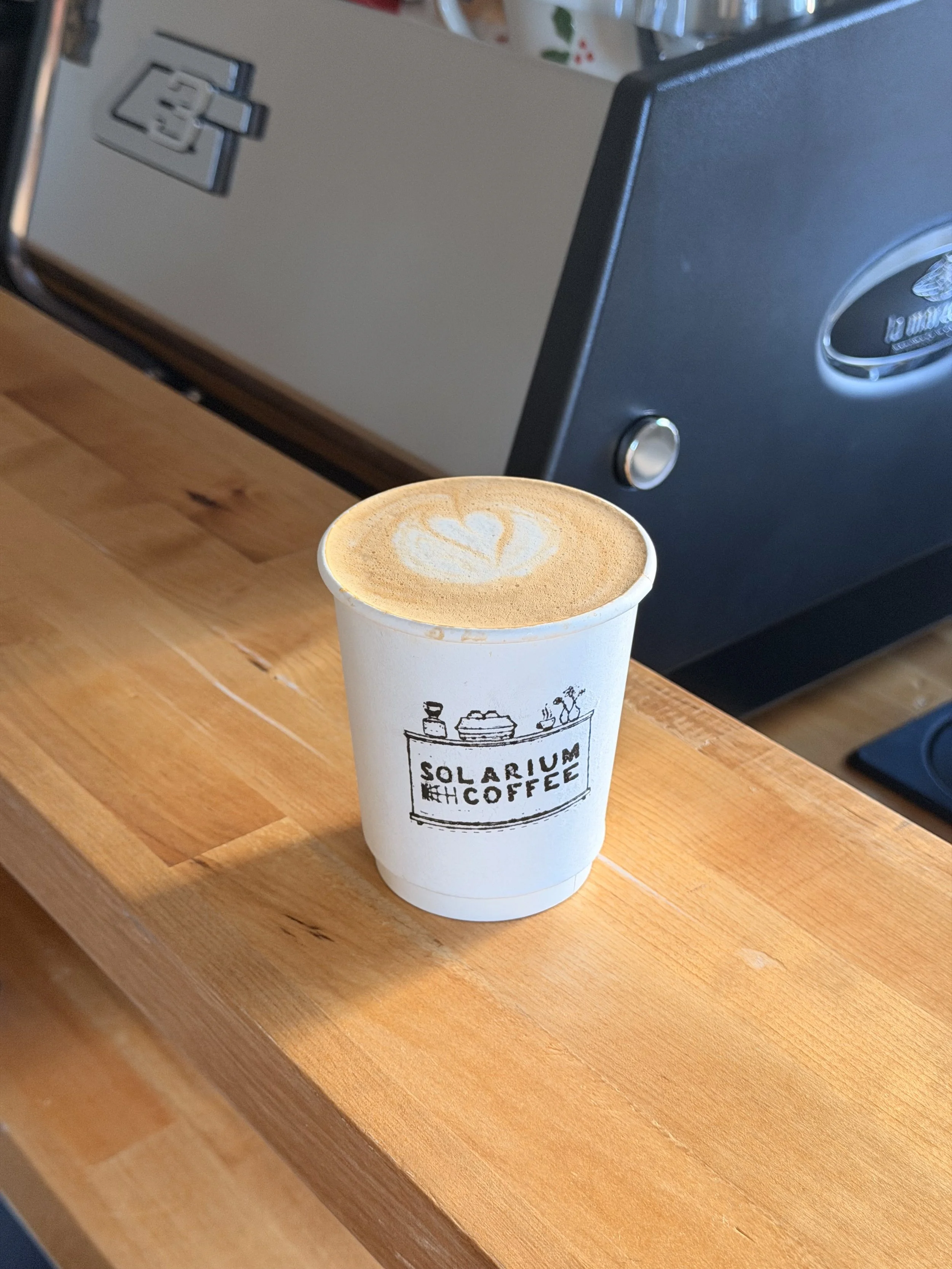 A cup of coffee with latte art on top, sitting on a wooden counter with a Solaris Coffee logo on the cup, in front of a coffee machine.