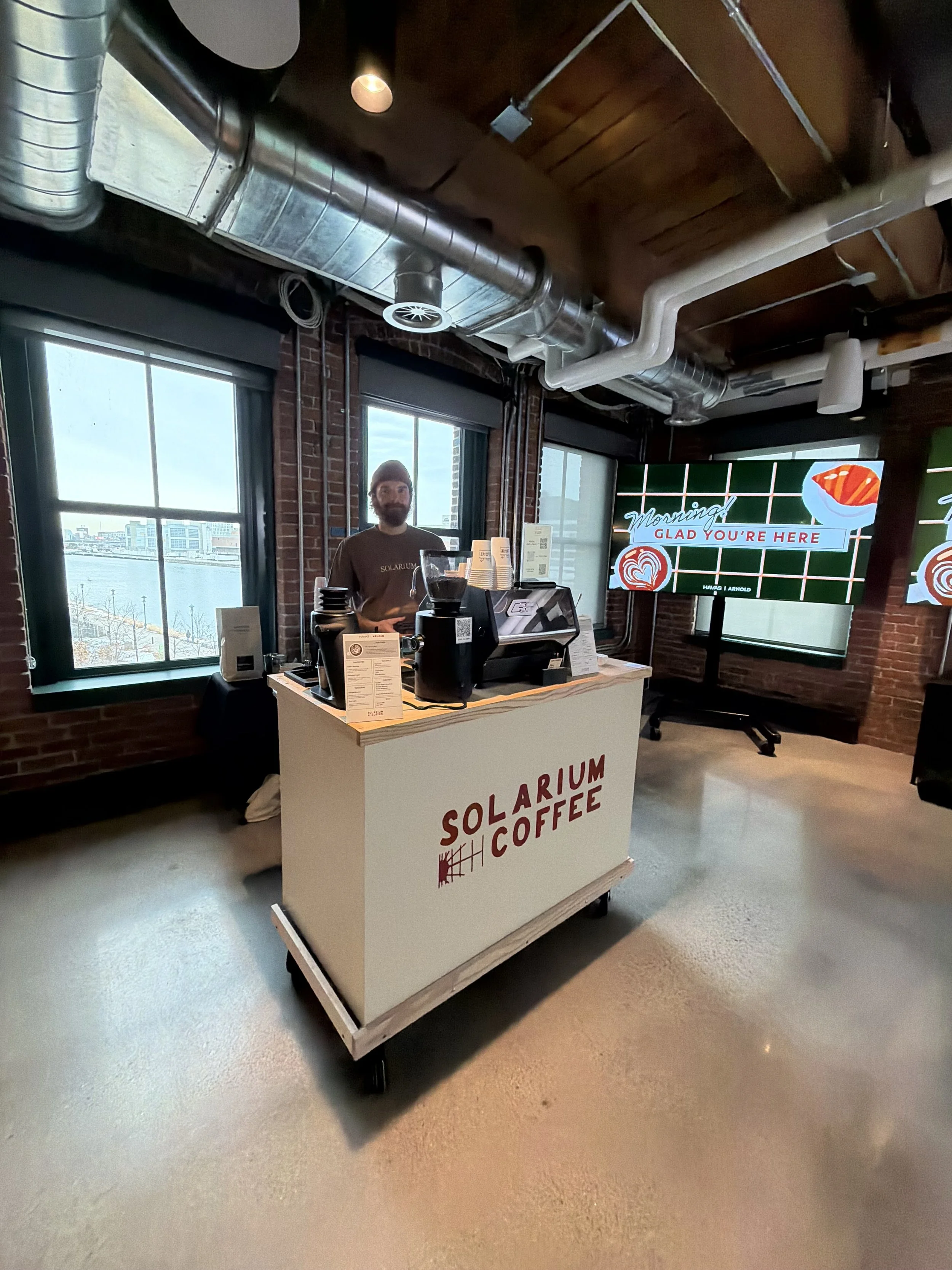 Indoor cafe with a barista at Solarium Coffee stand, large windows with cityscape view, digital screen with morning greeting, exposed brick and ductwork ceiling.