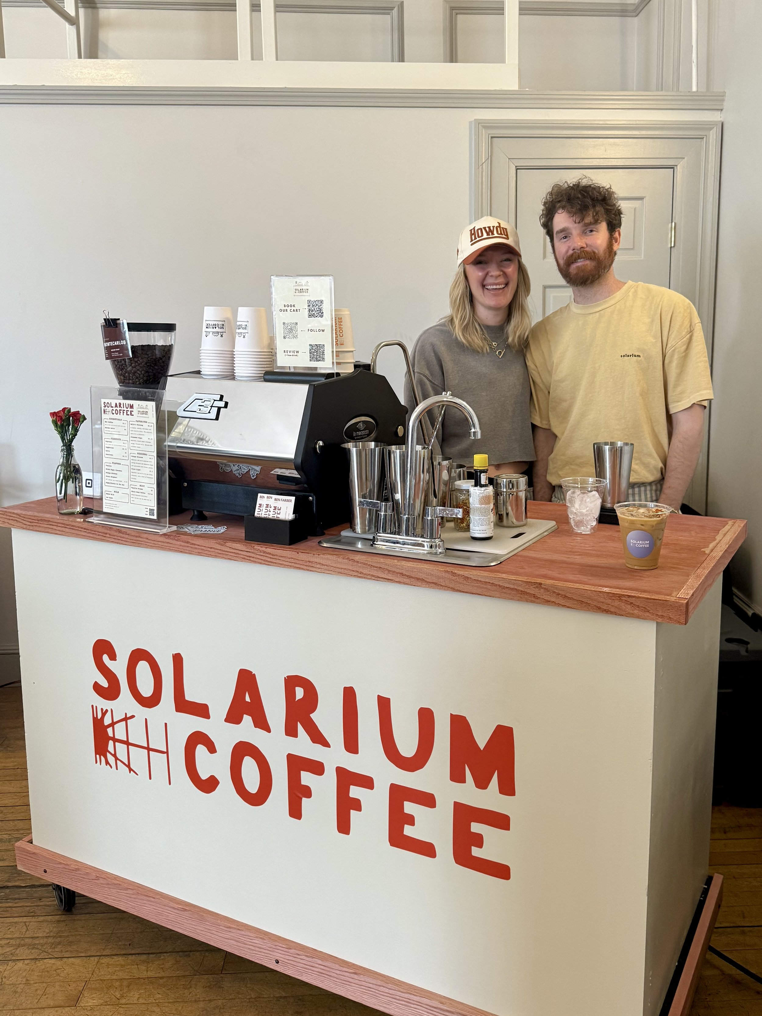 Two people standing behind a coffee cart labeled 'Solarium Coffee' inside a building. The woman is smiling and wearing a cap that says 'Howdy', and the man is smiling with a beard and wearing a yellow t-shirt. On the counter are coffee cups, a vase with a flower, and coffee-making supplies.