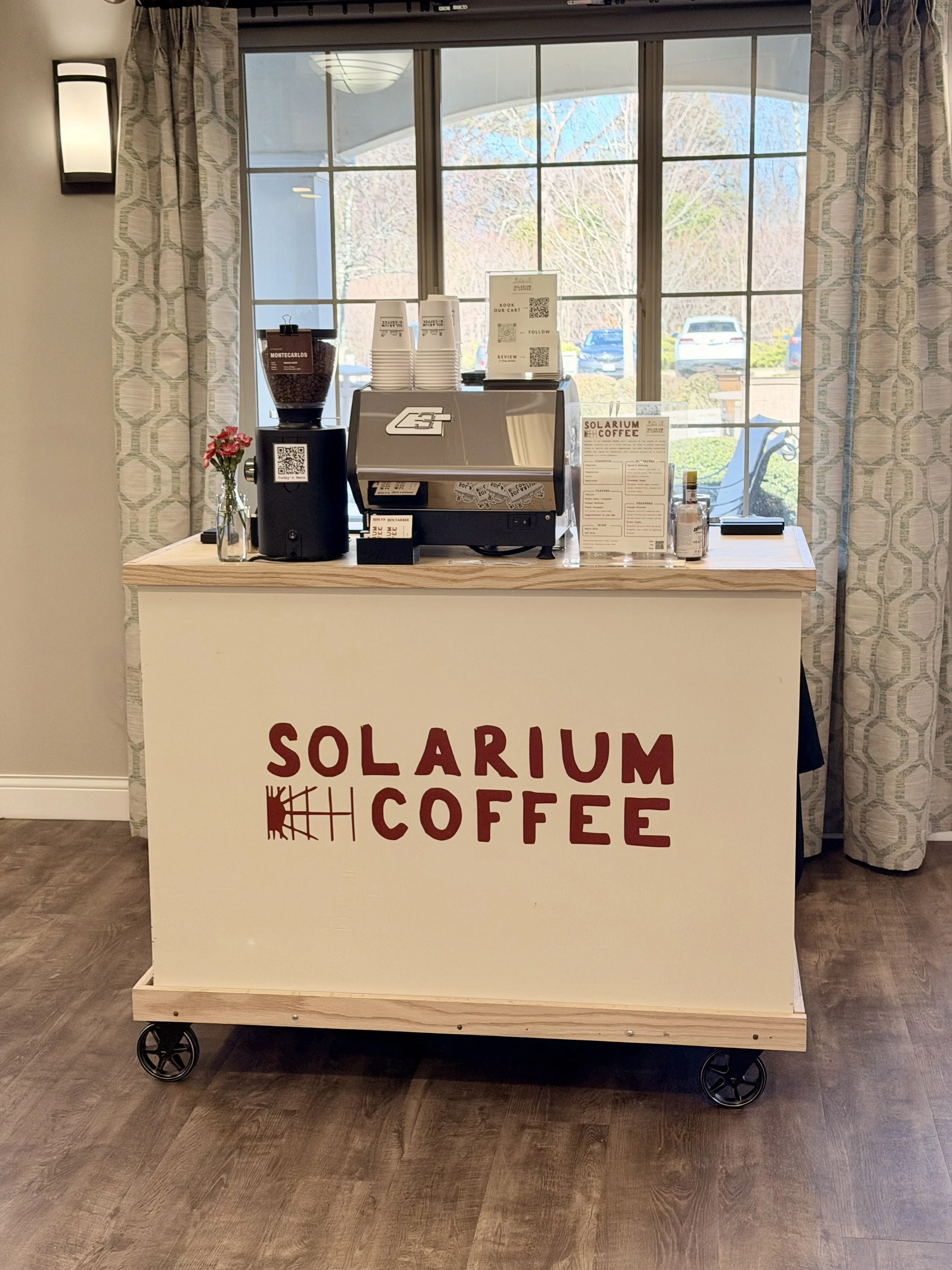 A coffee kiosk named Solarium Coffee with a large window behind it, a coffee machine, a small bouquet of pink flowers, a container of coffee beans, and a menu, all on a wooden cart with wheels.