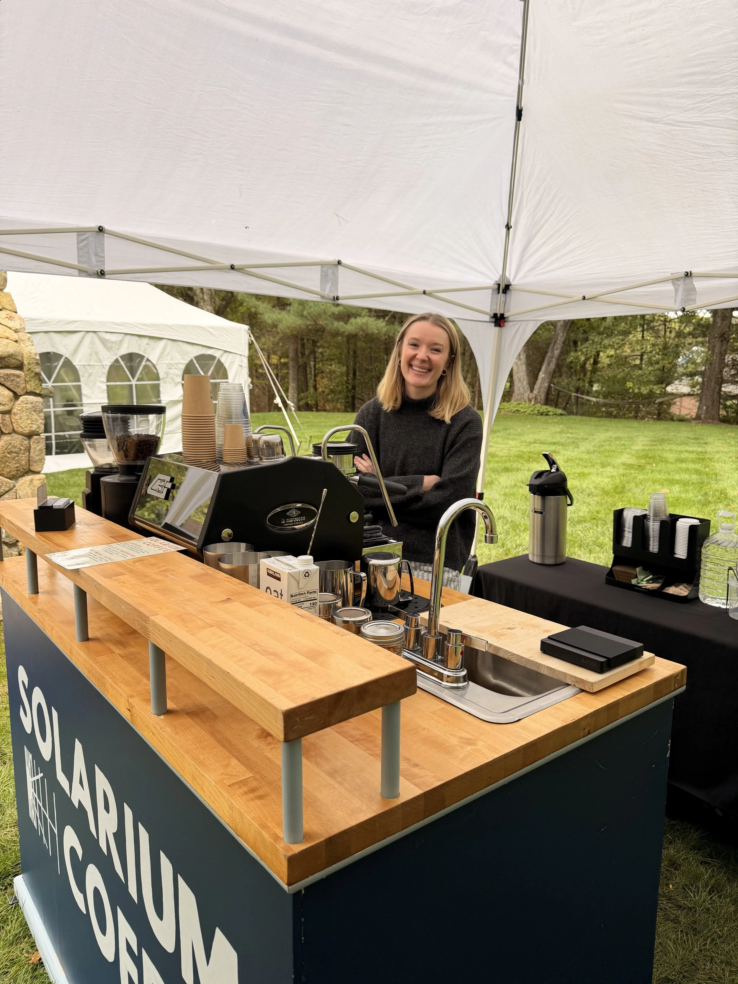 A smiling woman stands behind a coffee stand set up outdoors under a white canopy tent, with green grass and trees in the background, and a stone wall to the left.