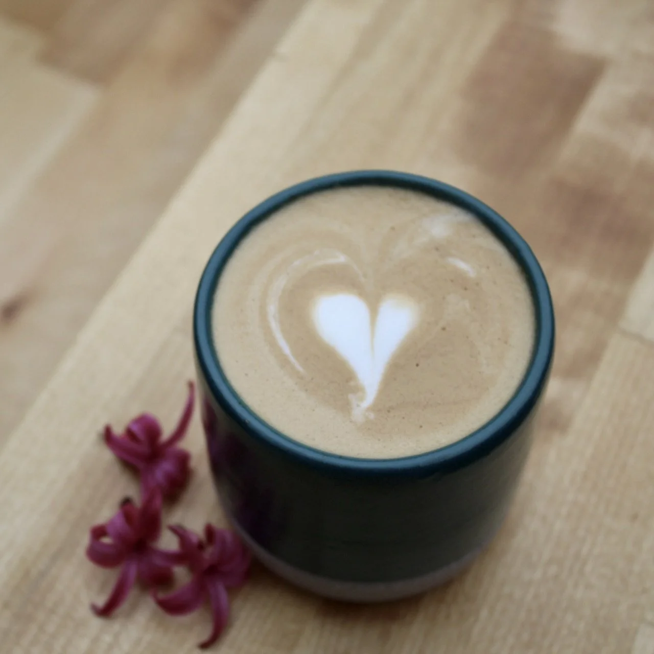 A cup of latte with heart-shaped foam art on a wooden table, with pink flowers beside the cup.