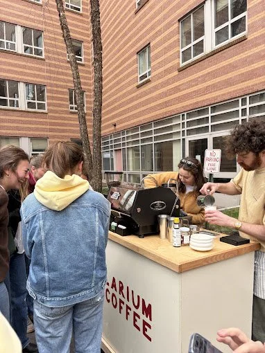 A group of people gathered around a coffee stand outside a brick building, with a man serving coffee.