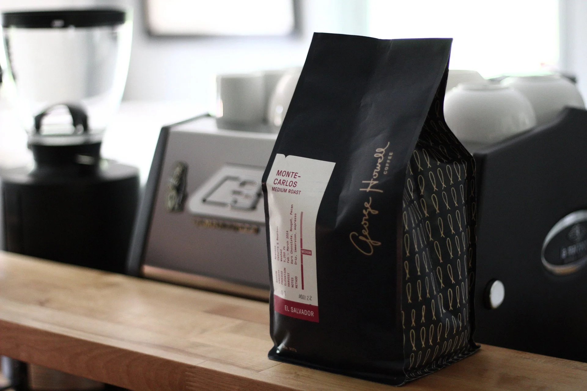 A black coffee bag labeled 'Espresso Hotel', 'Monte Carlos medium roast', with a label indicating it's from El Salvador, placed on a wooden countertop in a kitchen setting with a coffee machine, cups, and a container in the blurred background.