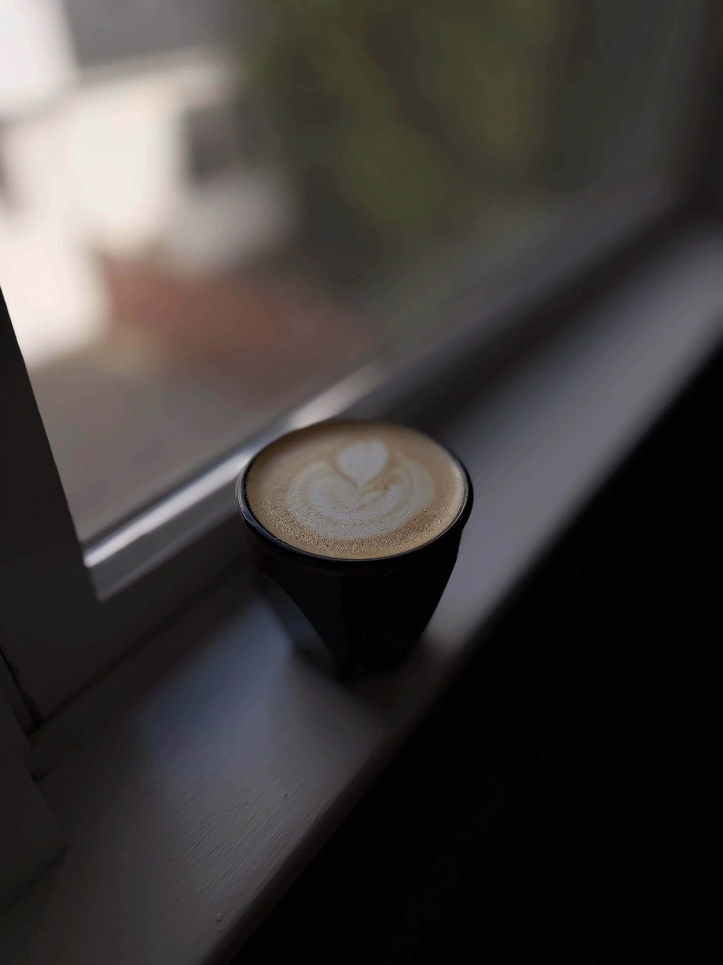 A cup of coffee with latte art sitting on a windowsill near a window.