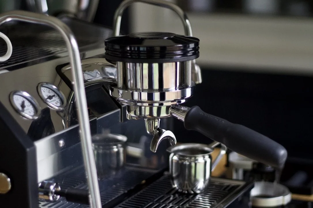 Close-up of a stainless steel espresso machine with a portafilter attached and a coffee cup positioned below the spout, with gauges and controls visible on the machine.