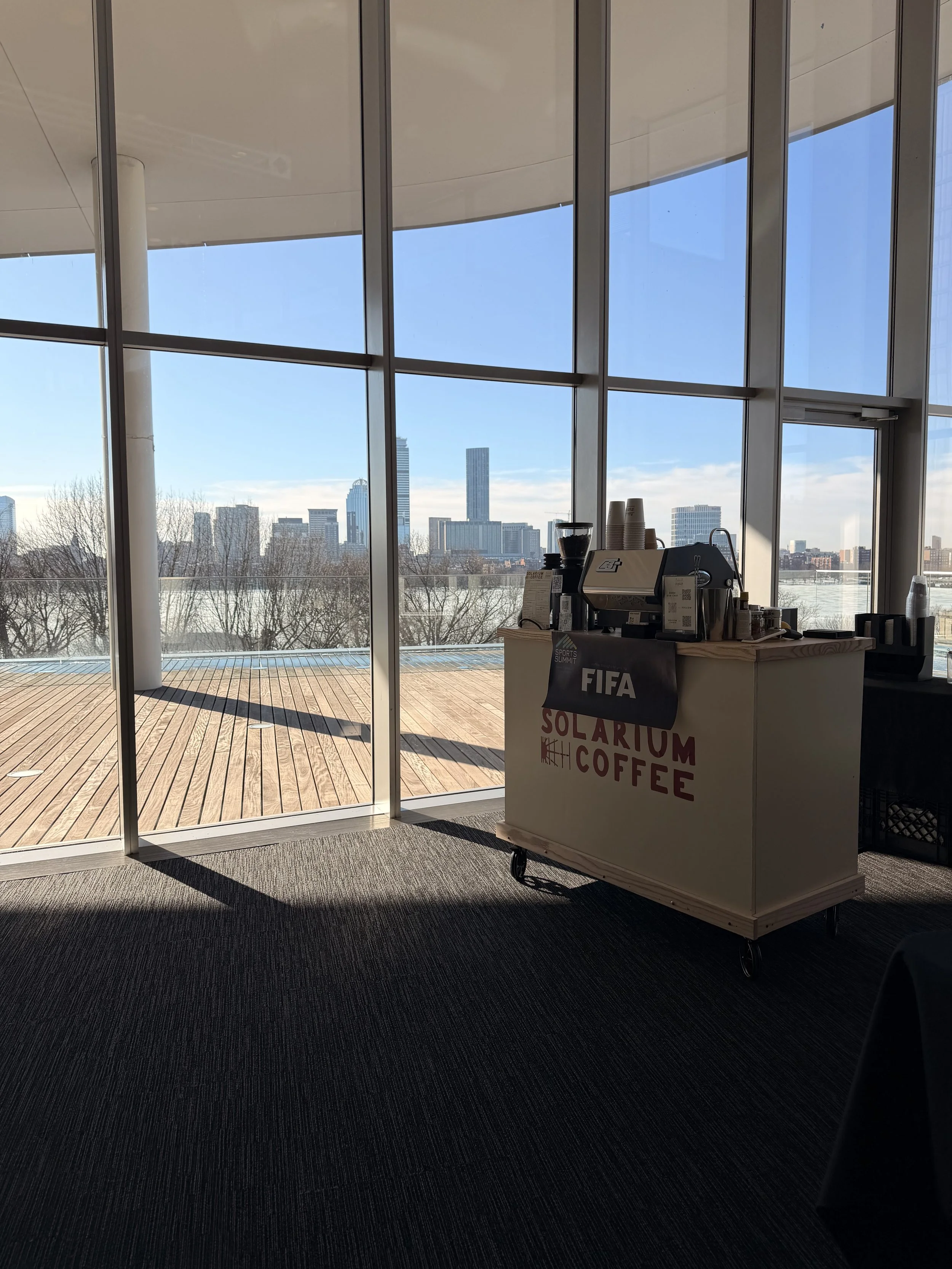 A coffee cart with a sign that says 'FIFA Solarium Coffee,' set up inside near large windows overlooking a cityscape with tall buildings, a clear sky, and leafless trees.