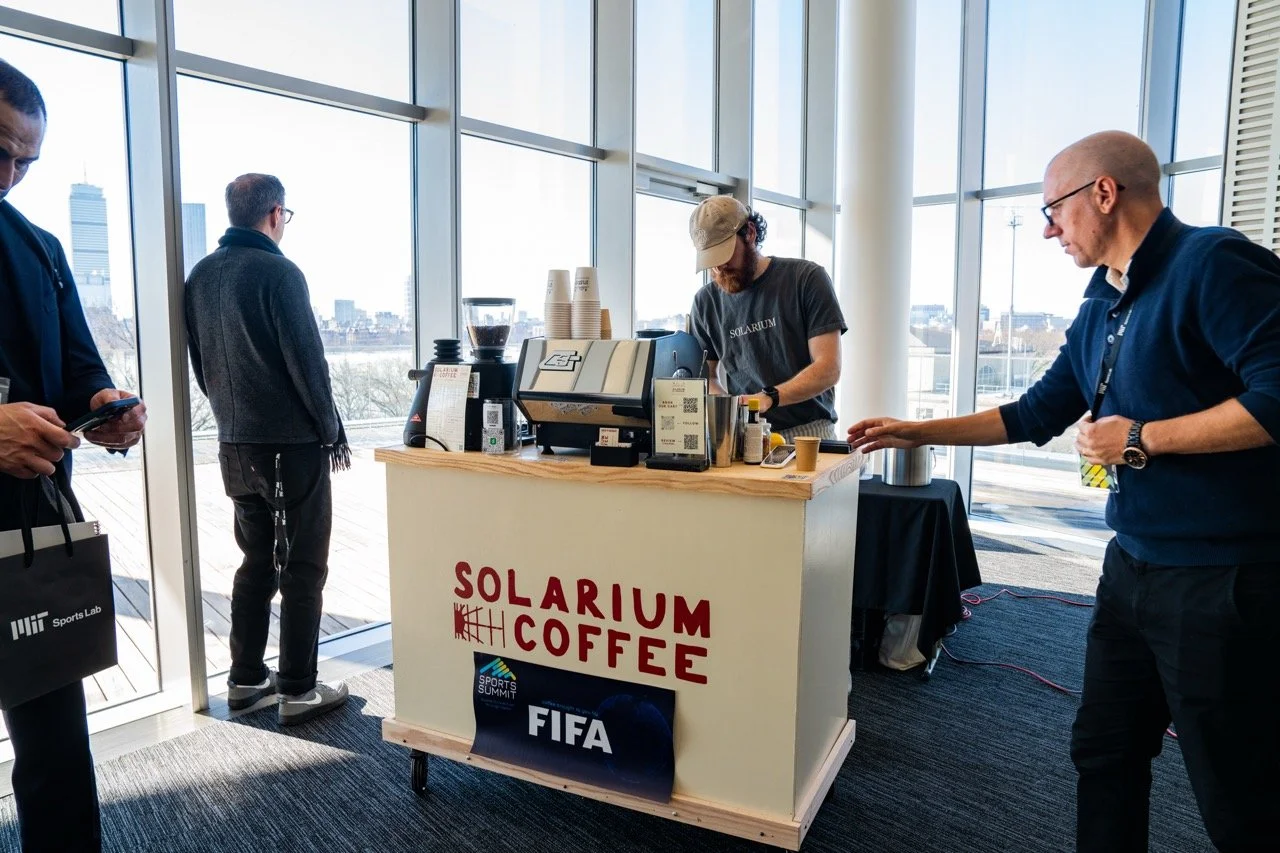 A coffee stand labeled 'SOLARIUM COFFEE' in a glass-walled room with three customers and one barista. Two customers are waiting in line, one using a smartphone, the other with hands in pockets. The barista is preparing coffee. The background shows a cityscape with buildings and a cloudy sky.