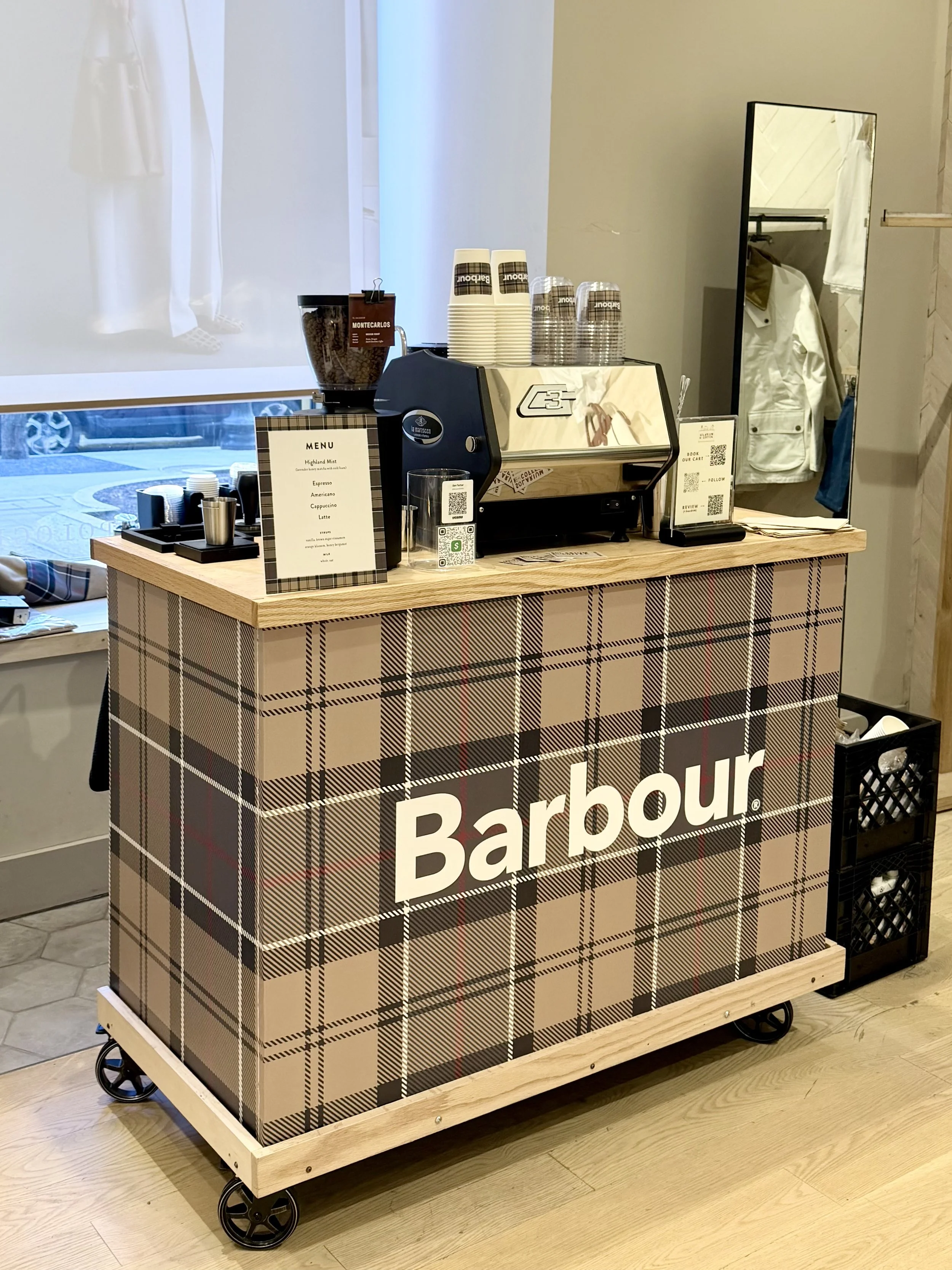 A mobile coffee cart with the word 'Barbour' on the front, featuring a plaid pattern and a wooden top, equipped with a coffee machine, cups, and coffee beans, inside a retail store.