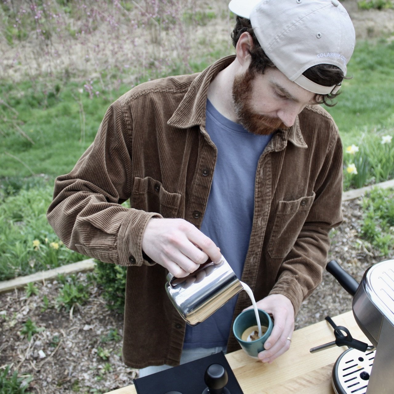 A man with a beard pouring steamed milk into a cup of coffee outdoors, with daffodils and greenery in the background.