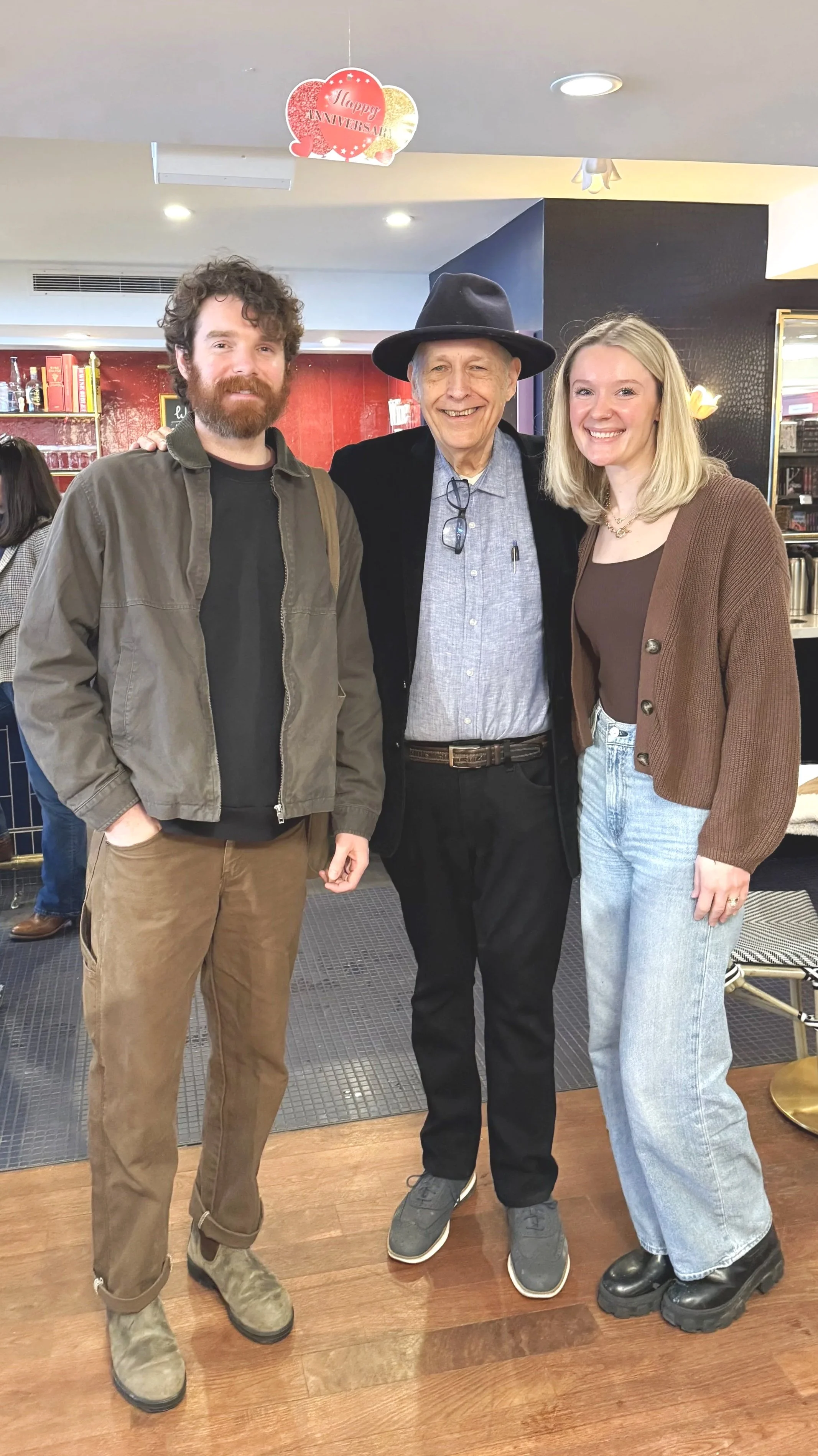 Three people standing together in a café or restaurant, smiling at the camera. The man in the center is wearing a black hat, glasses hanging from his shirt, and a black jacket. The man on the left has curly hair, a beard, and is dressed in a brown jacket and pants. The woman on the right has blonde hair, a brown cardigan, and light-wash jeans.