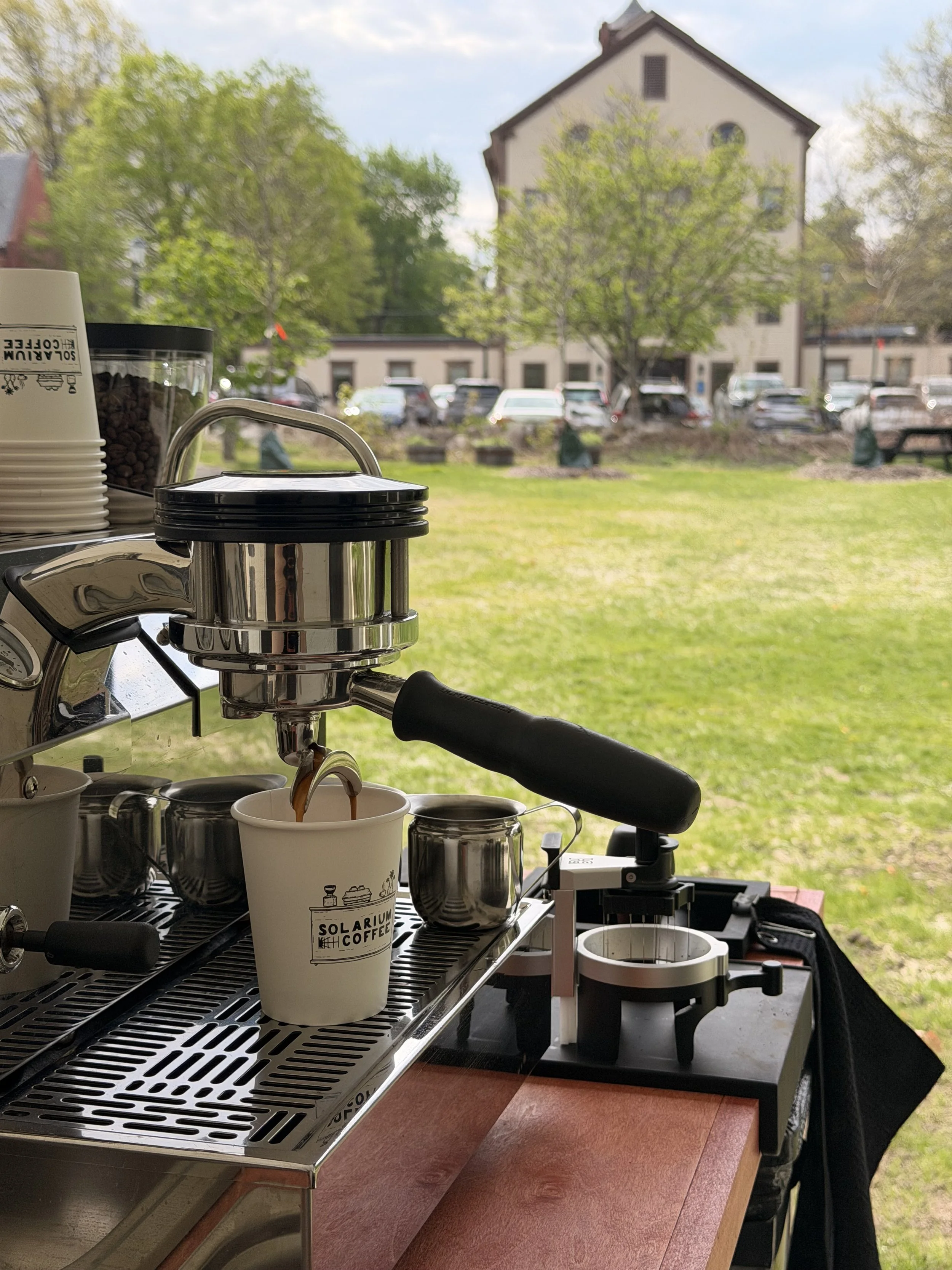 A coffee cart set up outdoors with a coffee machine in focus, pouring coffee into a paper cup with the logo 'Solarium Coffee'. Background shows trees, parked cars, and a building.