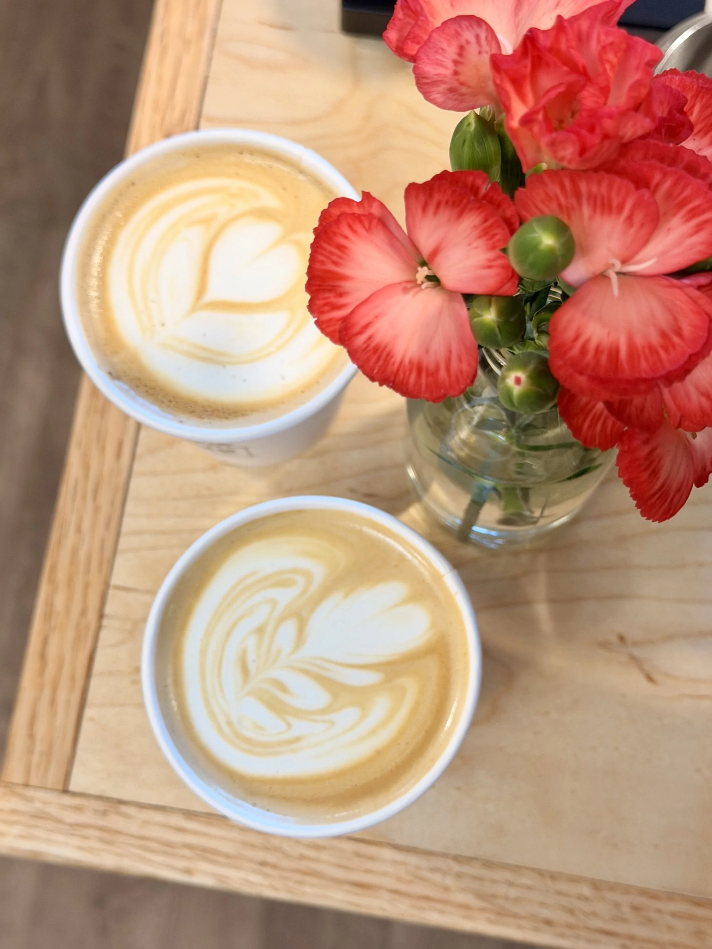 Two cups of coffee with latte art, a glass vase with pink and red flowers on a wooden table.