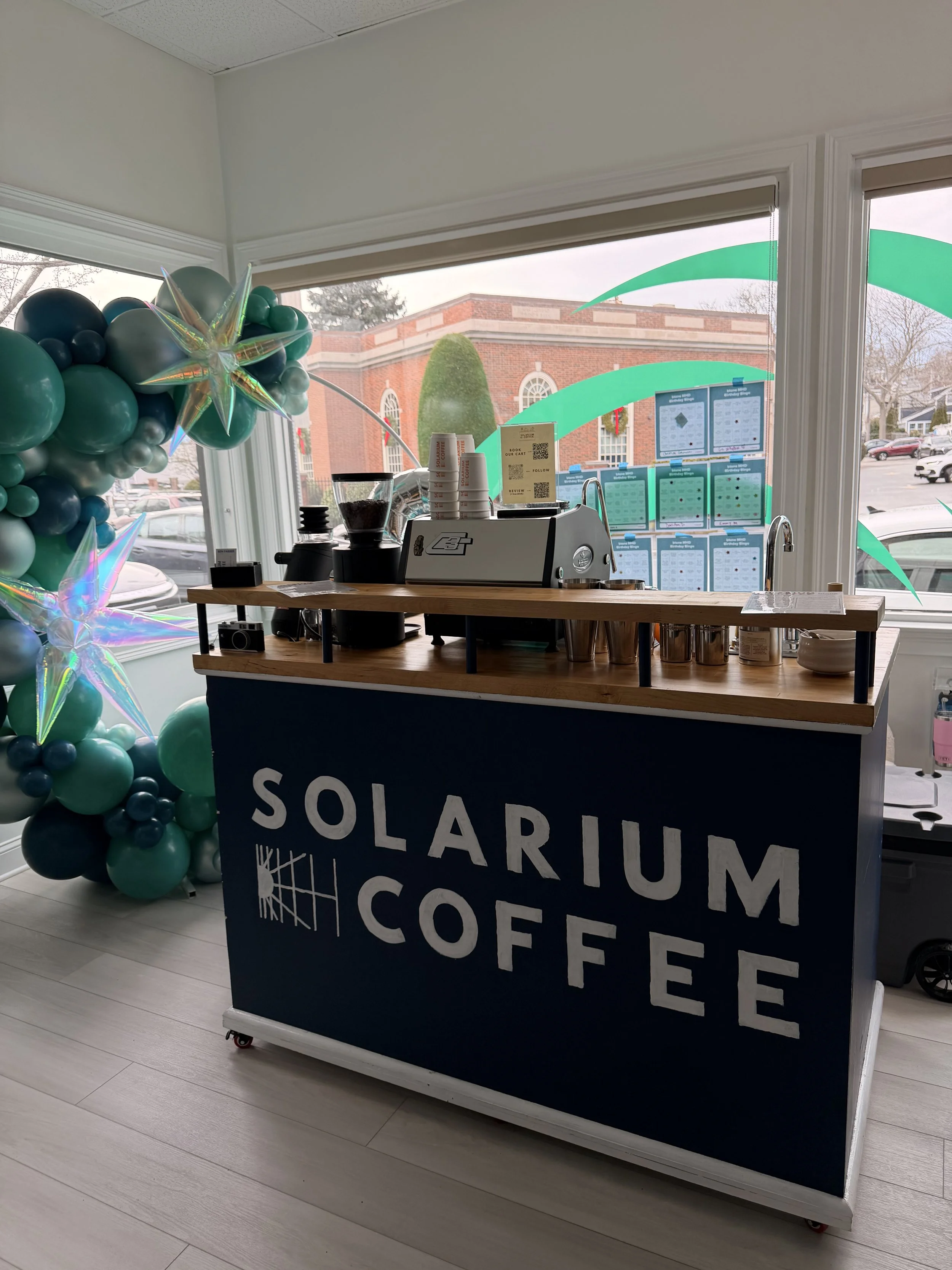A coffee stand named 'Solarium Coffee' with a balloon decoration and a wooden countertop, inside a bright room with large windows showing a brick building outside.