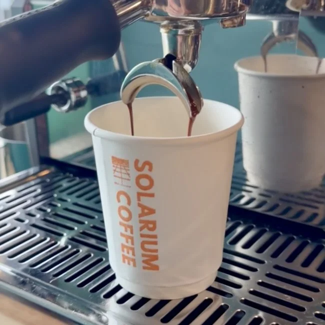 A coffee machine dispensing coffee into a white paper cup labeled 'SOLARIUM COFFEE' on a metal drip tray.