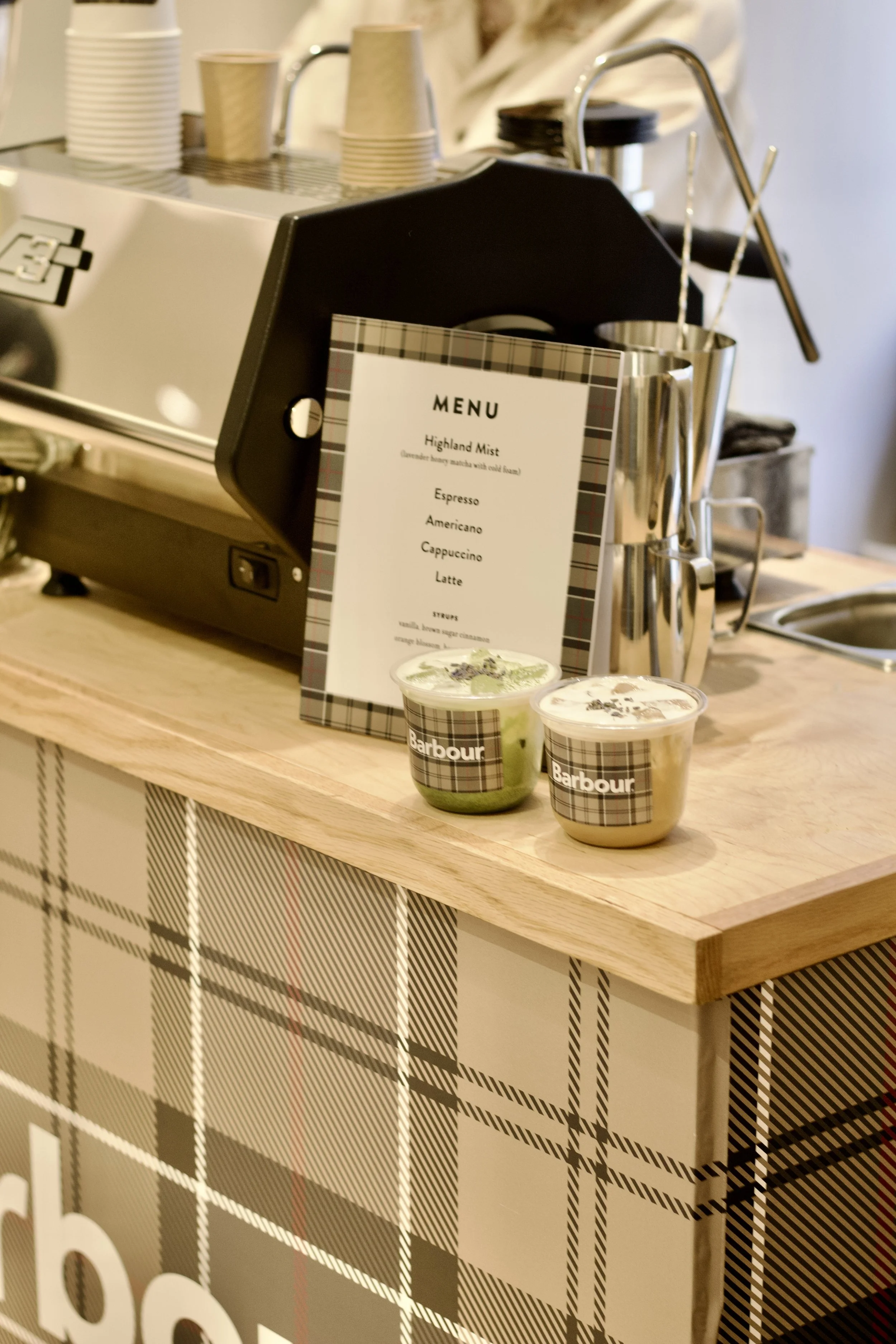 Two cups of coffee on a wooden counter, a menu card displaying coffee options, and a coffee machine in the background.
